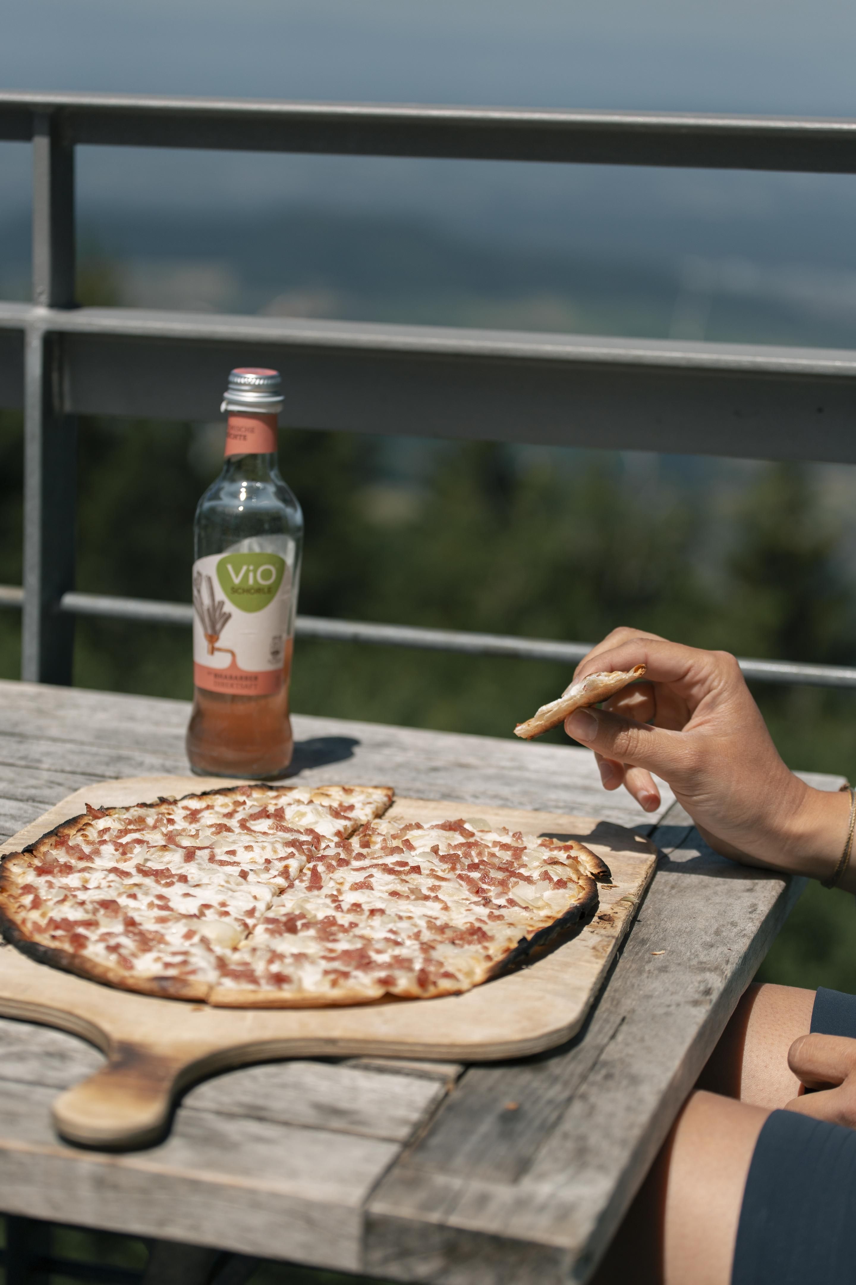 Flammkuchen auf einem Holztisch mit einer Hand, die ein Stück hält. Im Hintergrund eine Flasche Vio Schorle und Aussicht vom Schauinsland.