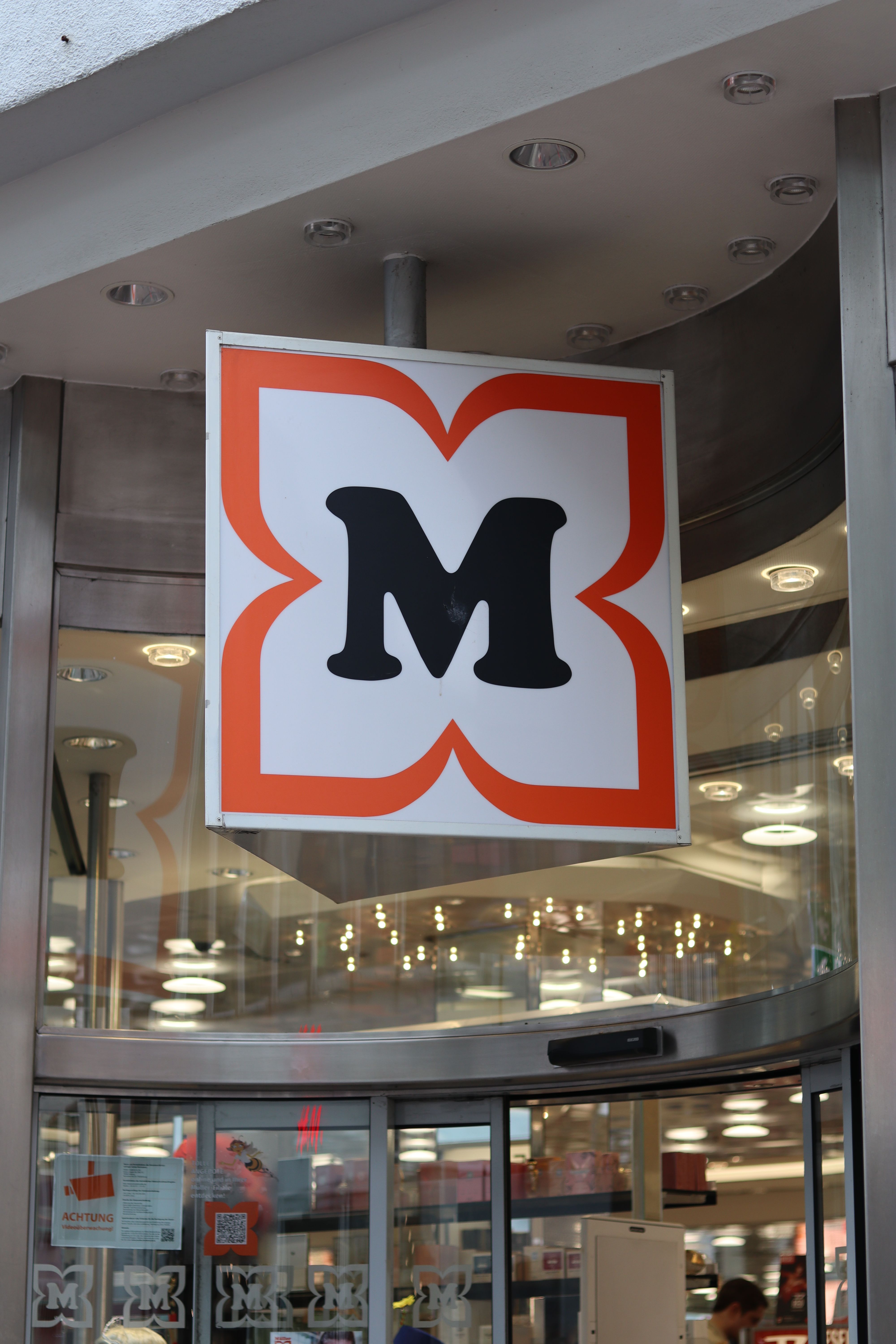 Eingang eines Müller Drogeriemarkts mit dem markanten M-Logo über der Tür. Innen sind Regale und Beleuchtung zu sehen.
