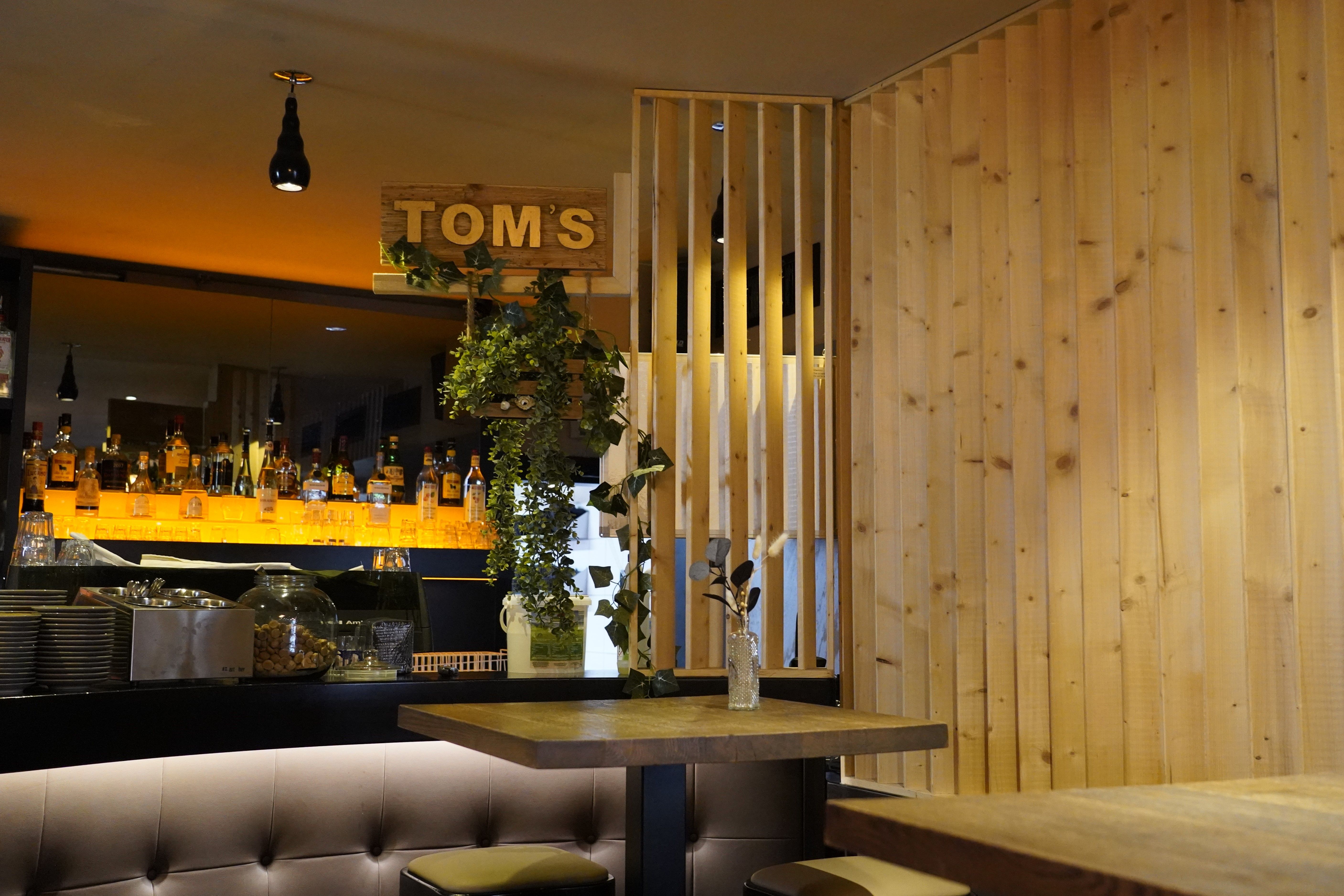 Gemütliches Restaurant mit Holzwänden, Bar im Hintergrund, beleuchtetem Regal mit Flaschen und einem Schild 'Tom's'. Pflanzen schmücken den Raum.
