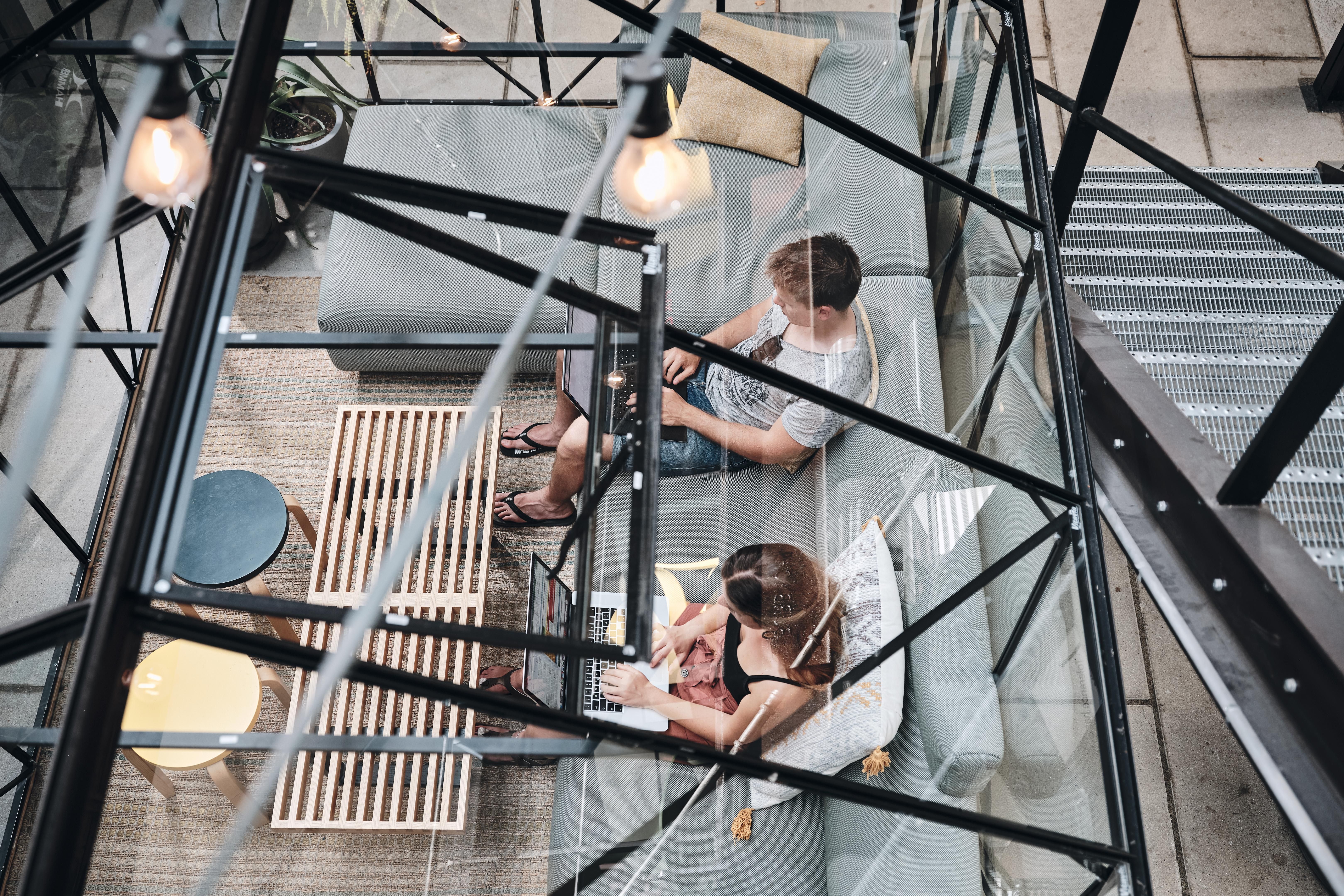 Zwei Personen sitzen in einem verglasten Raum auf Sofas und arbeiten an Laptops. Der Raum ist modern eingerichtet mit Teppich und kleinen Tischen.