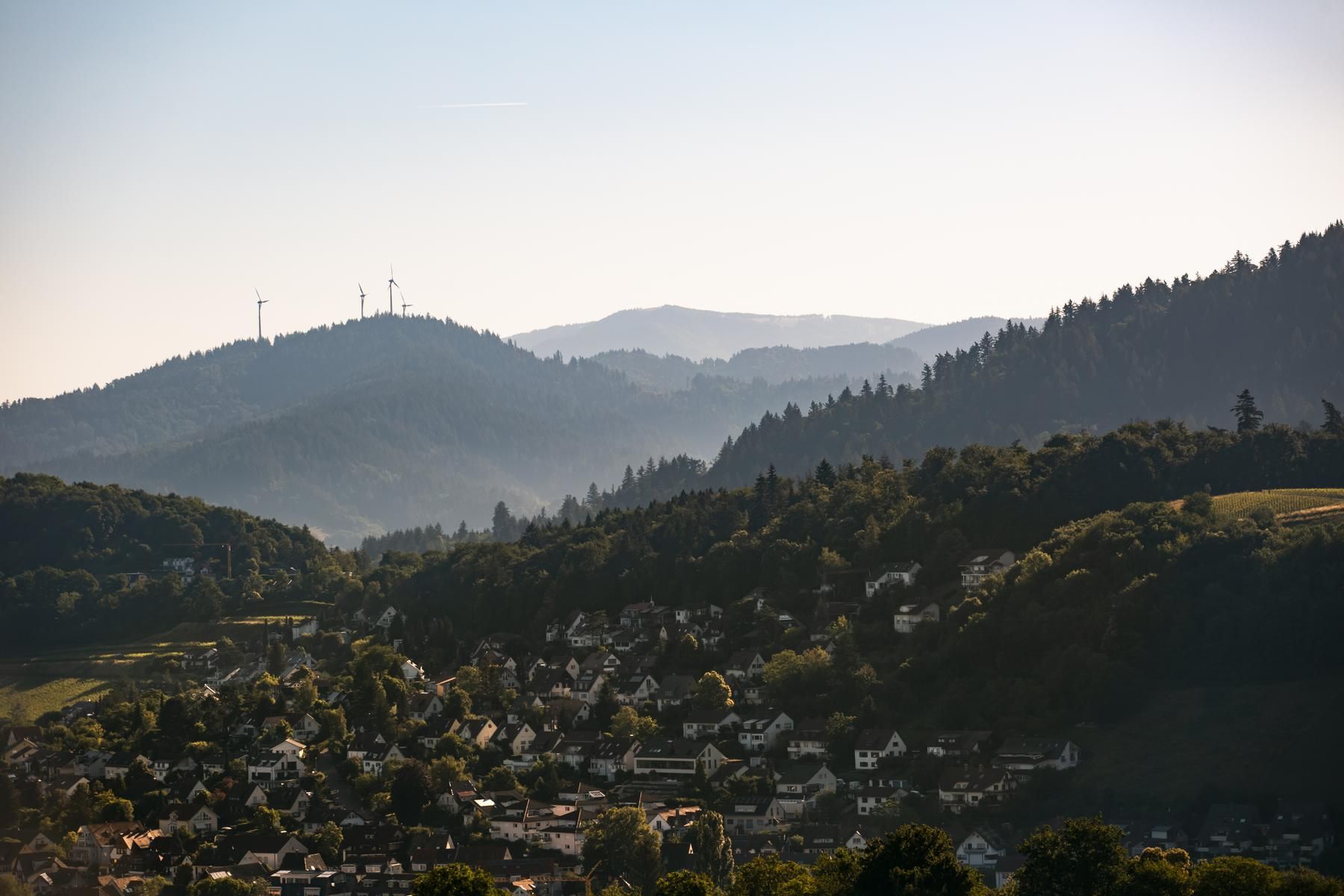 Panoramablick auf den Rosskopf bei Freiburg mit Windrädern auf einem bewaldeten Hügel und einer Siedlung im Vordergrund.