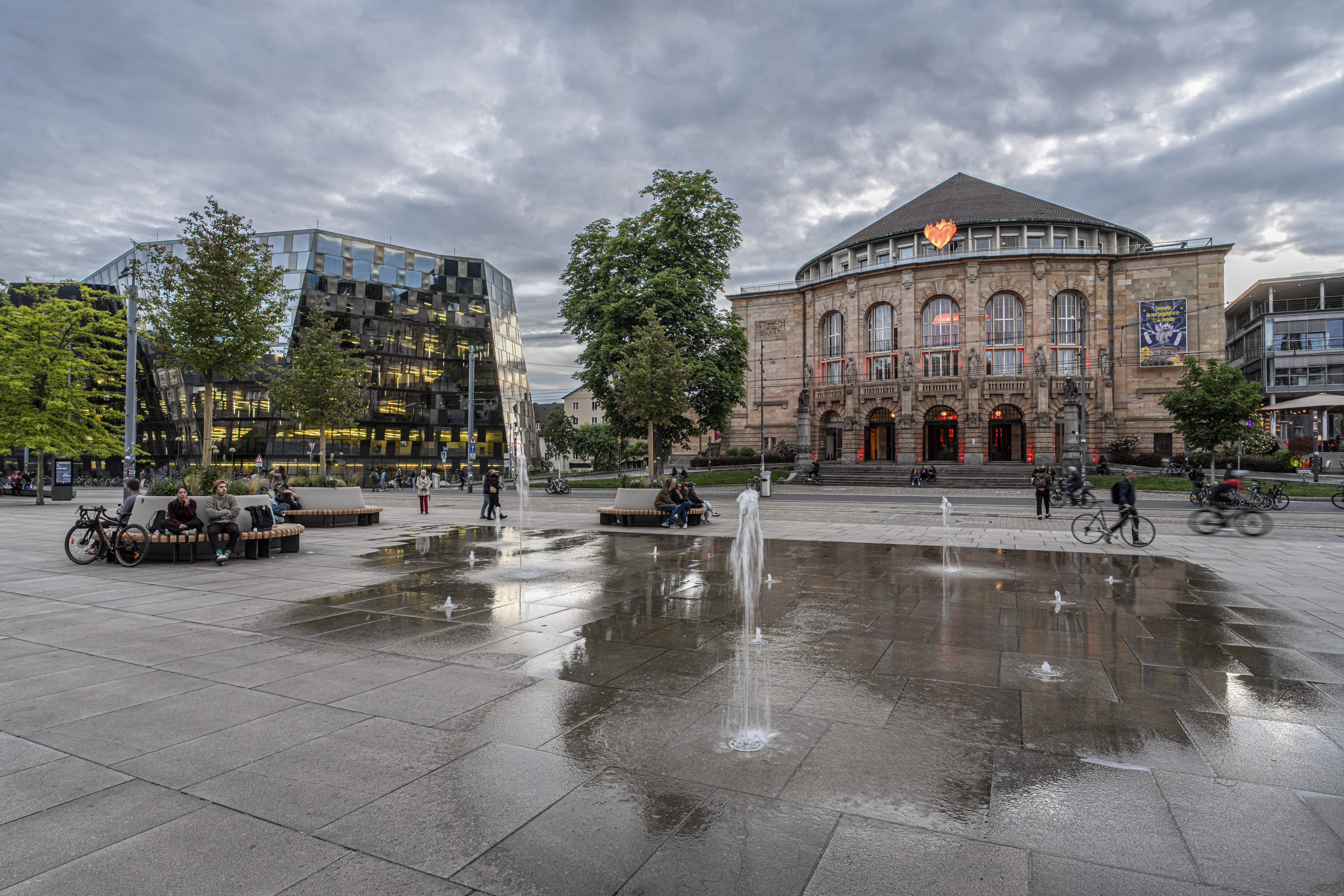 Das Freiburger Stadttheater bei bewölktem Himmel, umgeben von modernen Gebäuden und einem Platz mit Wasserspielen.