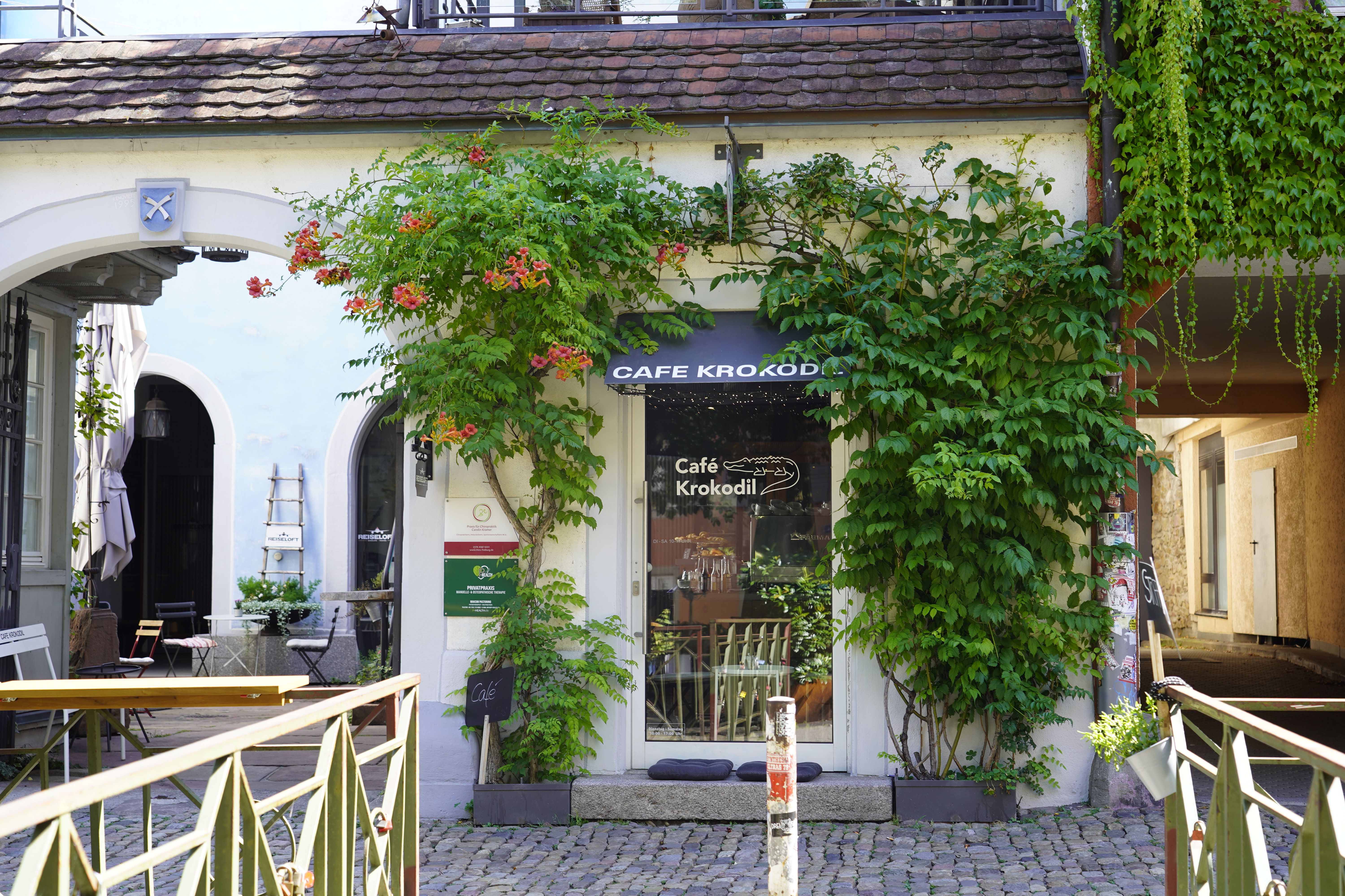 Das Café Krokodil mit Pflanzen an der Fassade, Kopfsteinpflaster und einem kleinen Außenbereich. Einladende Atmosphäre in einer ruhigen Gasse.