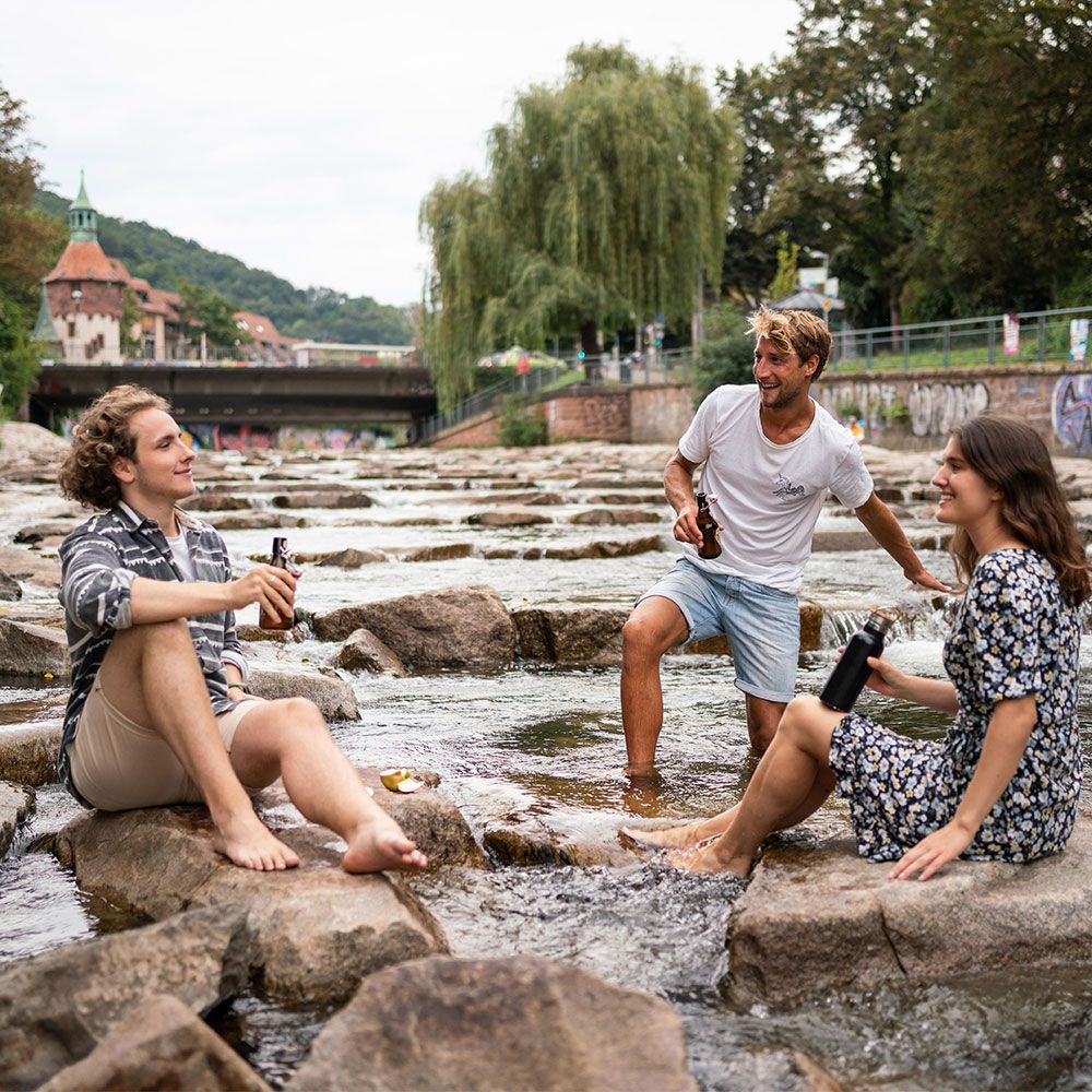 Freiburg im Sommer- Gruppe von 3 Menschen sitzt auf Steinen in der Dreisam 