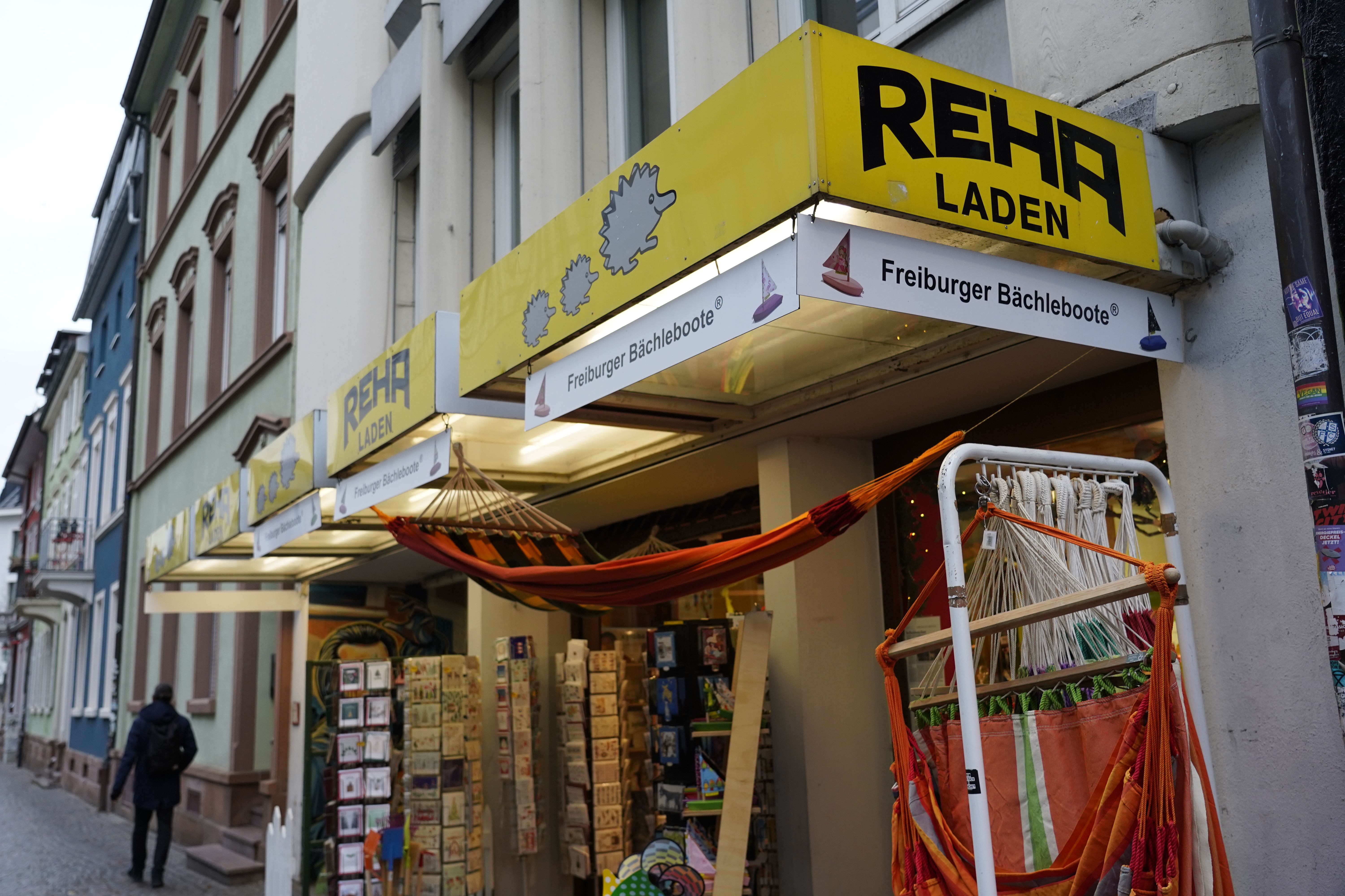 Ein Laden in Freiburg mit gelbem Schild 'Reha Laden'. Vor dem Geschäft hängen bunte Hängematten. Passanten gehen auf der Straße vorbei.