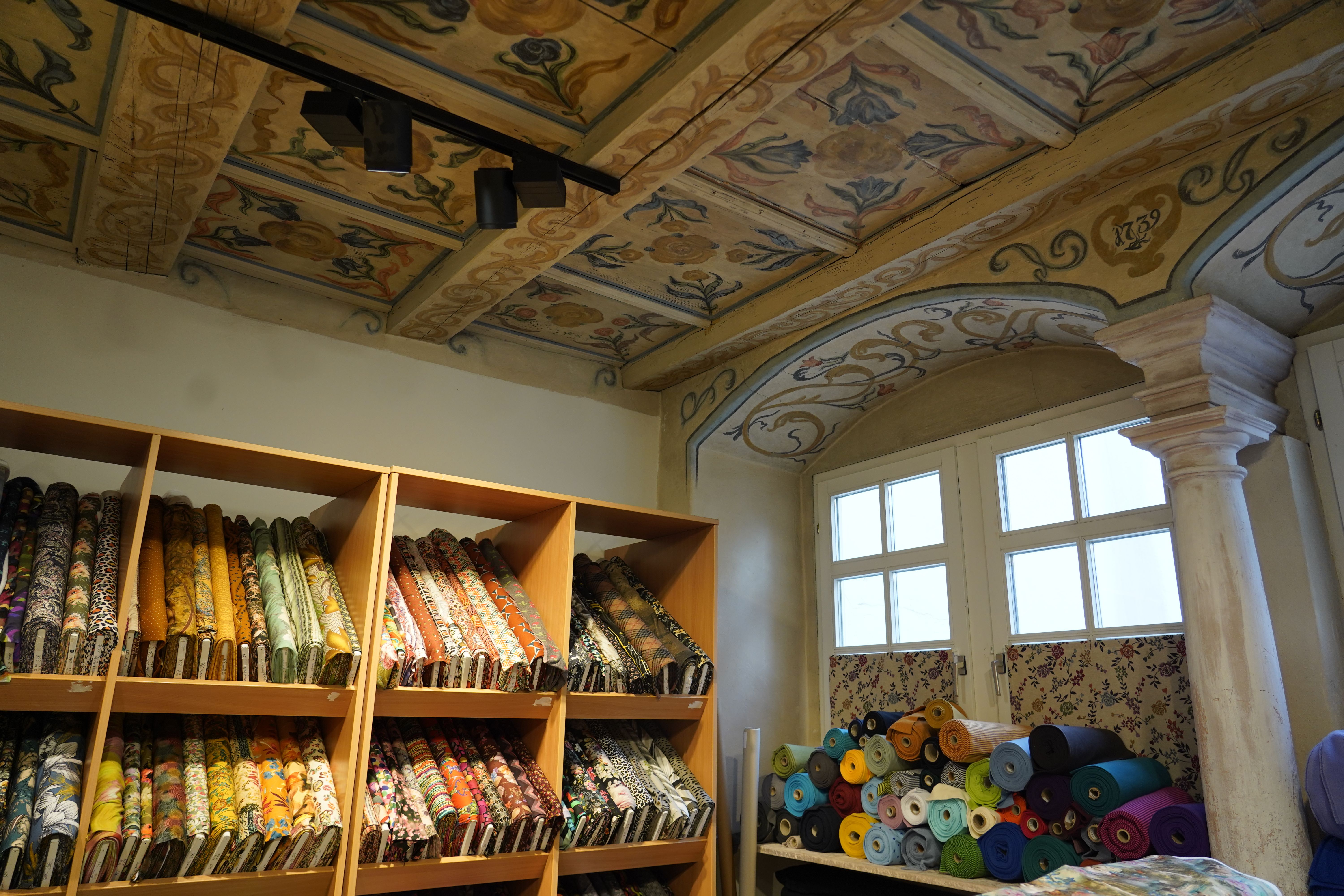 Ein Stoffladen mit kunstvoll bemalter Decke und Regalen voller bunter Stoffrollen. Eine Säule und ein Fenster sind ebenfalls sichtbar.