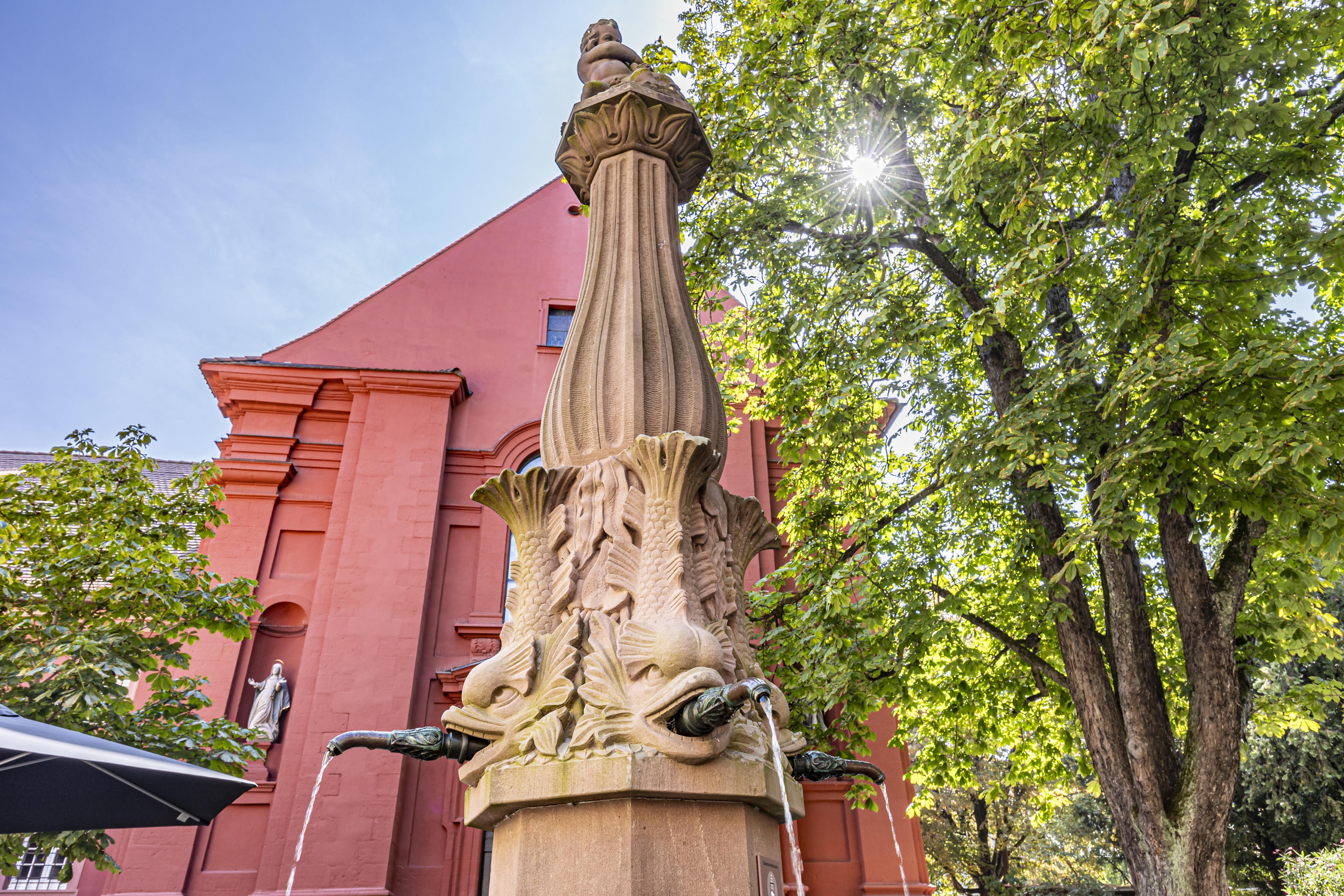 Ein kunstvoll verzierter Brunnen vor einem roten Gebäude in Freiburg, umgeben von Bäumen. Die Sonne scheint durch die Blätter.
