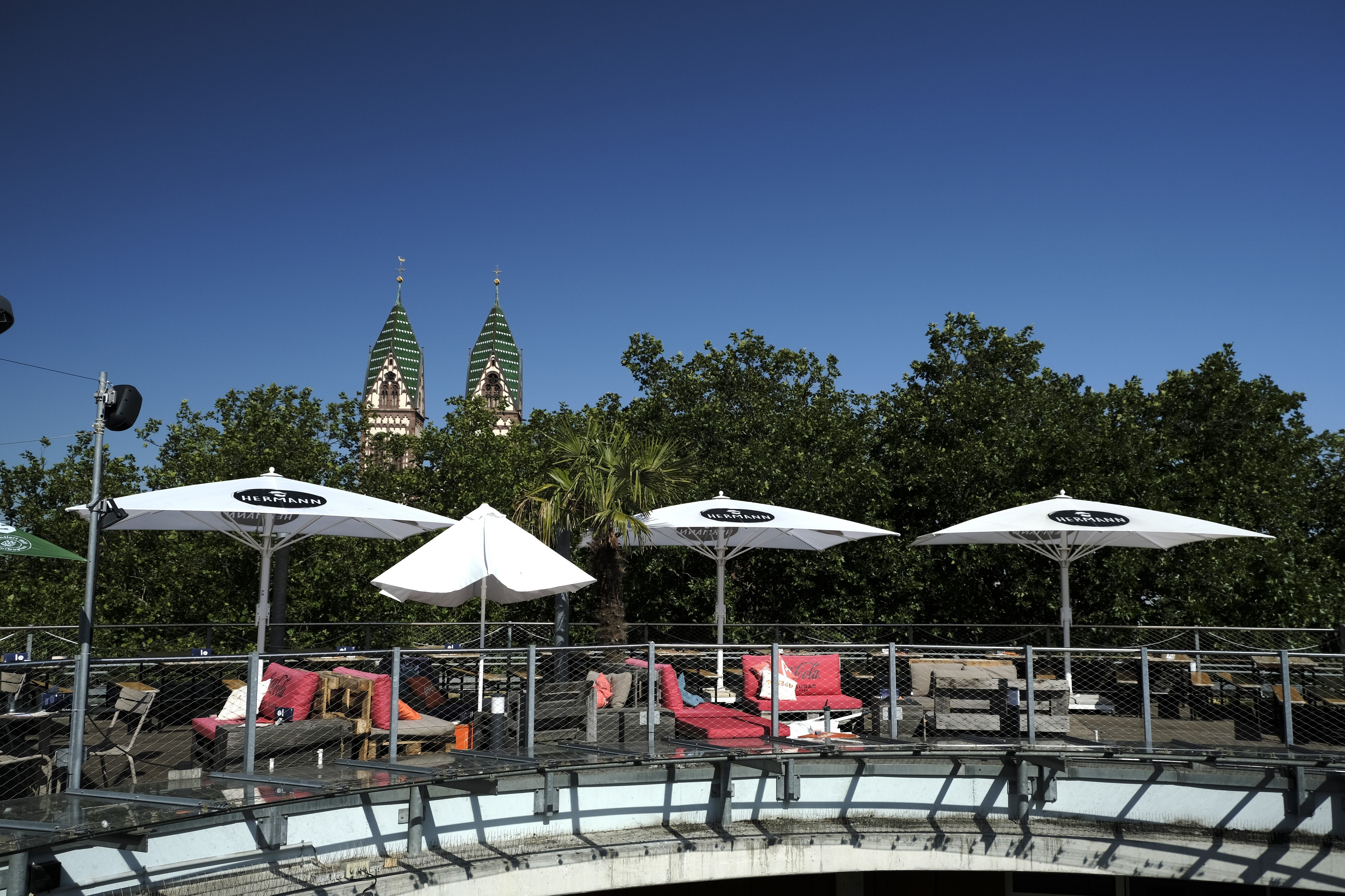 Terrasse des Café Hermann mit weißen Sonnenschirmen und Sitzgelegenheiten, umgeben von Bäumen. Im Hintergrund sind Kirchtürme zu sehen.