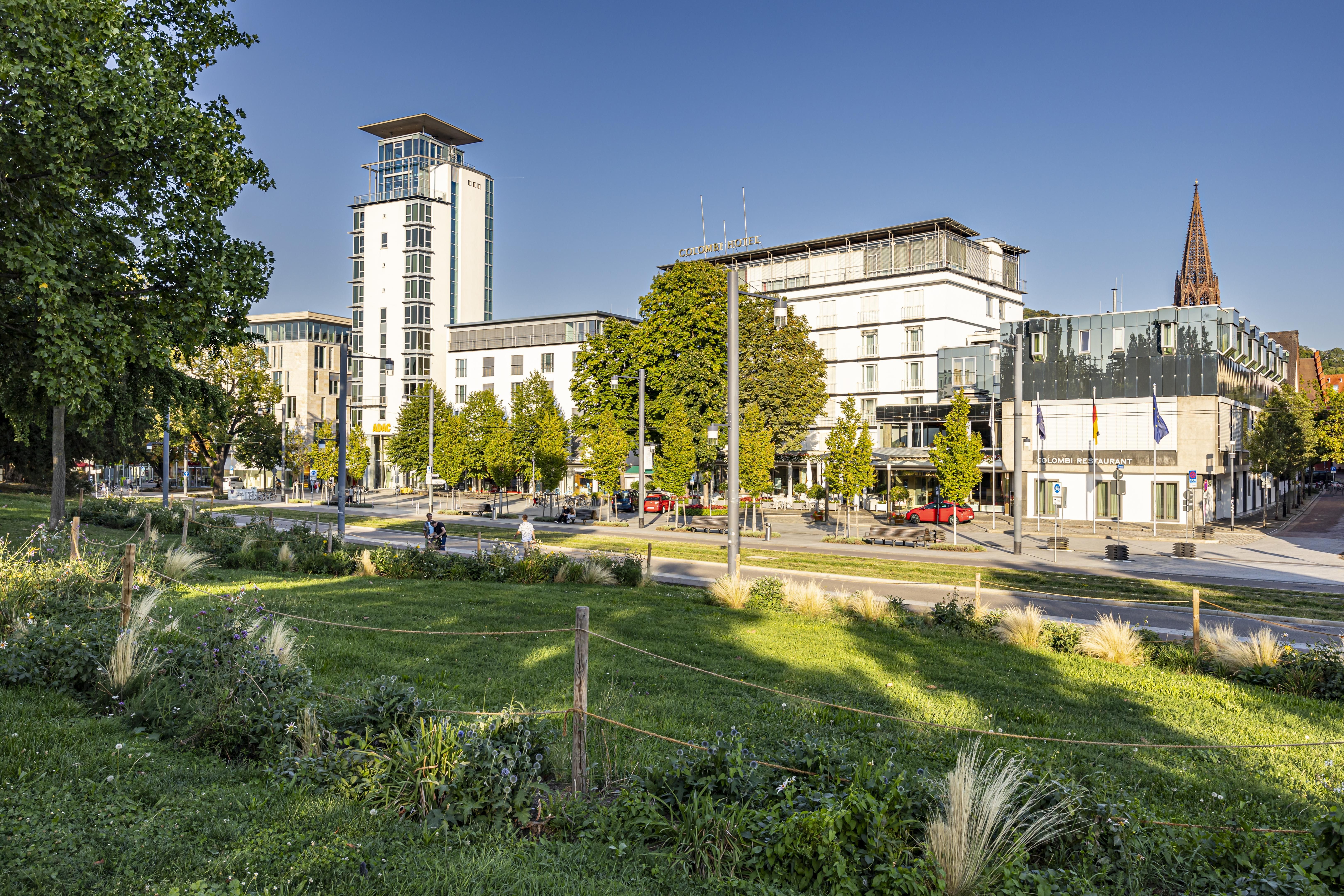 Blick auf das Colombi Hotel in Freiburg, umgeben von Bäumen und einem grünen Park. Im Hintergrund ist ein Kirchturm sichtbar.