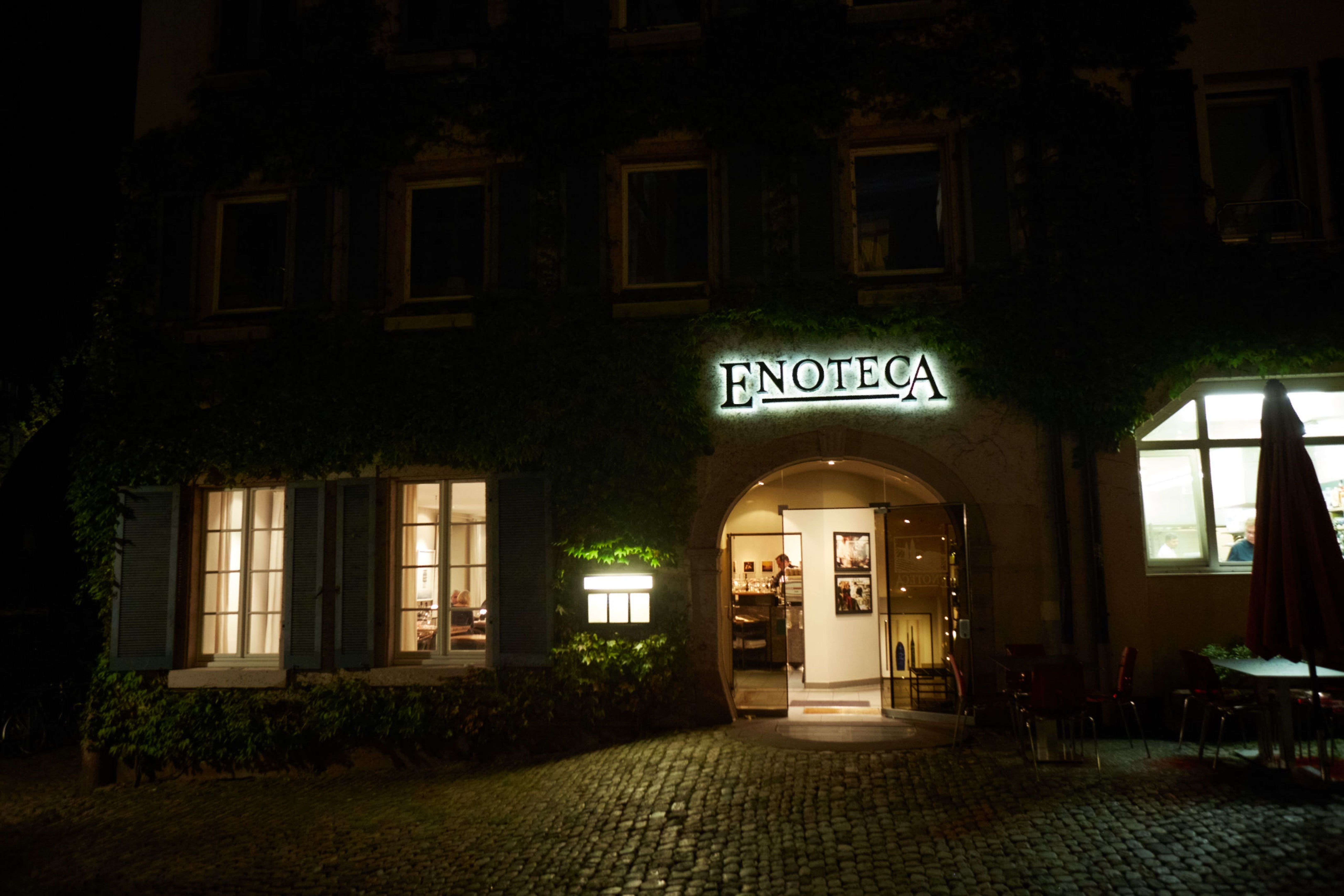 Beleuchteter Eingang der Enoteca in Freiburg bei Nacht, umgeben von Pflanzen und Kopfsteinpflaster.