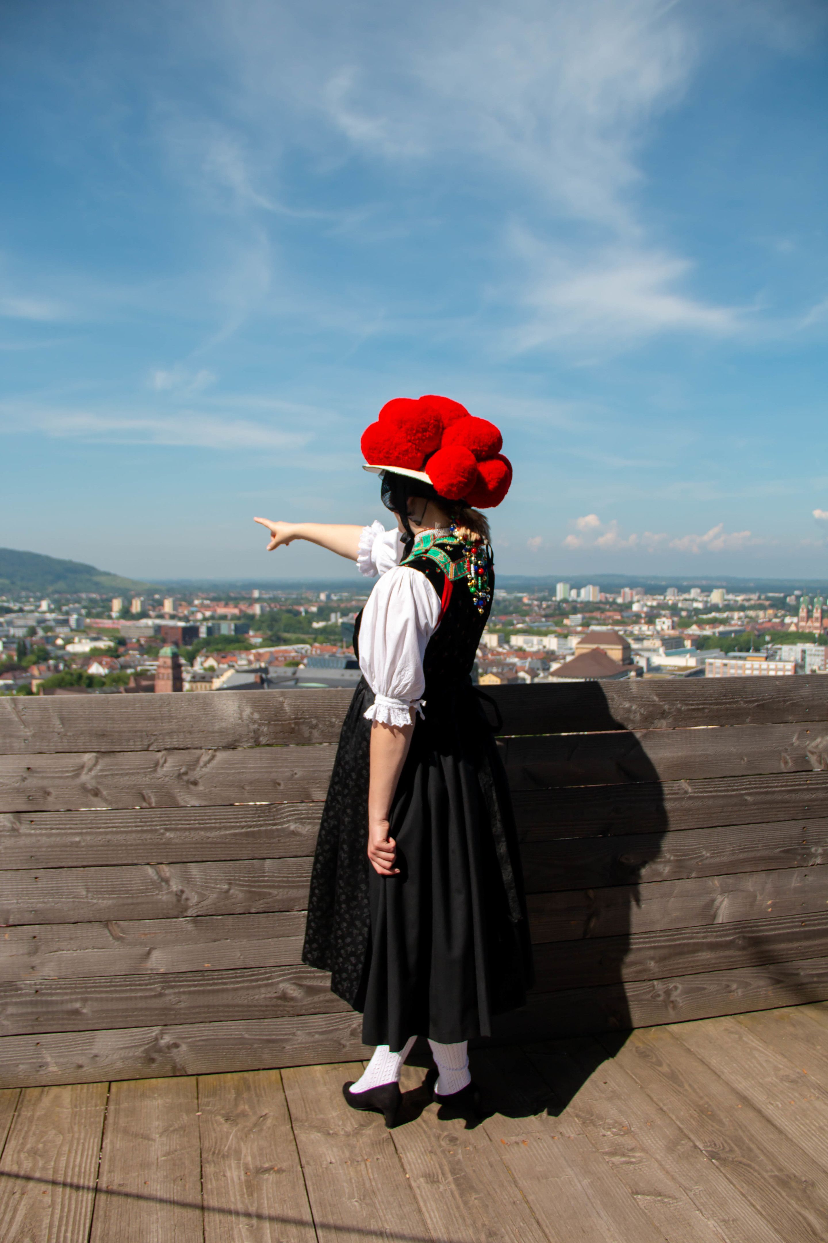 Eine Person in traditioneller Tracht mit rotem Bollenhut zeigt auf die Stadt Freiburg im Hintergrund. Der Himmel ist blau und klar.
