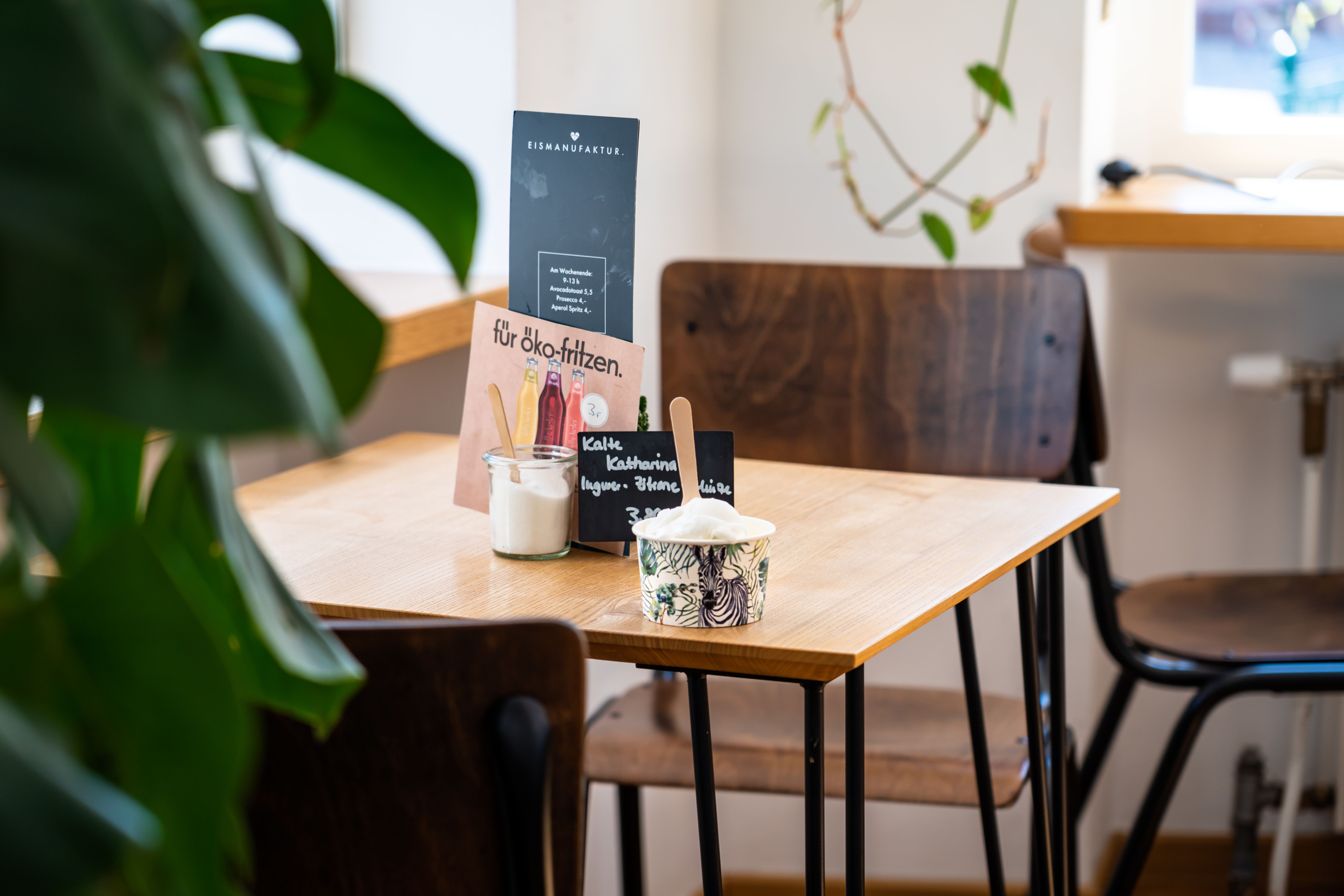 Ein Tisch in einem Café mit einem Eisbecher. Im Hintergrund sind Holzstühle und eine Pflanze zu sehen.