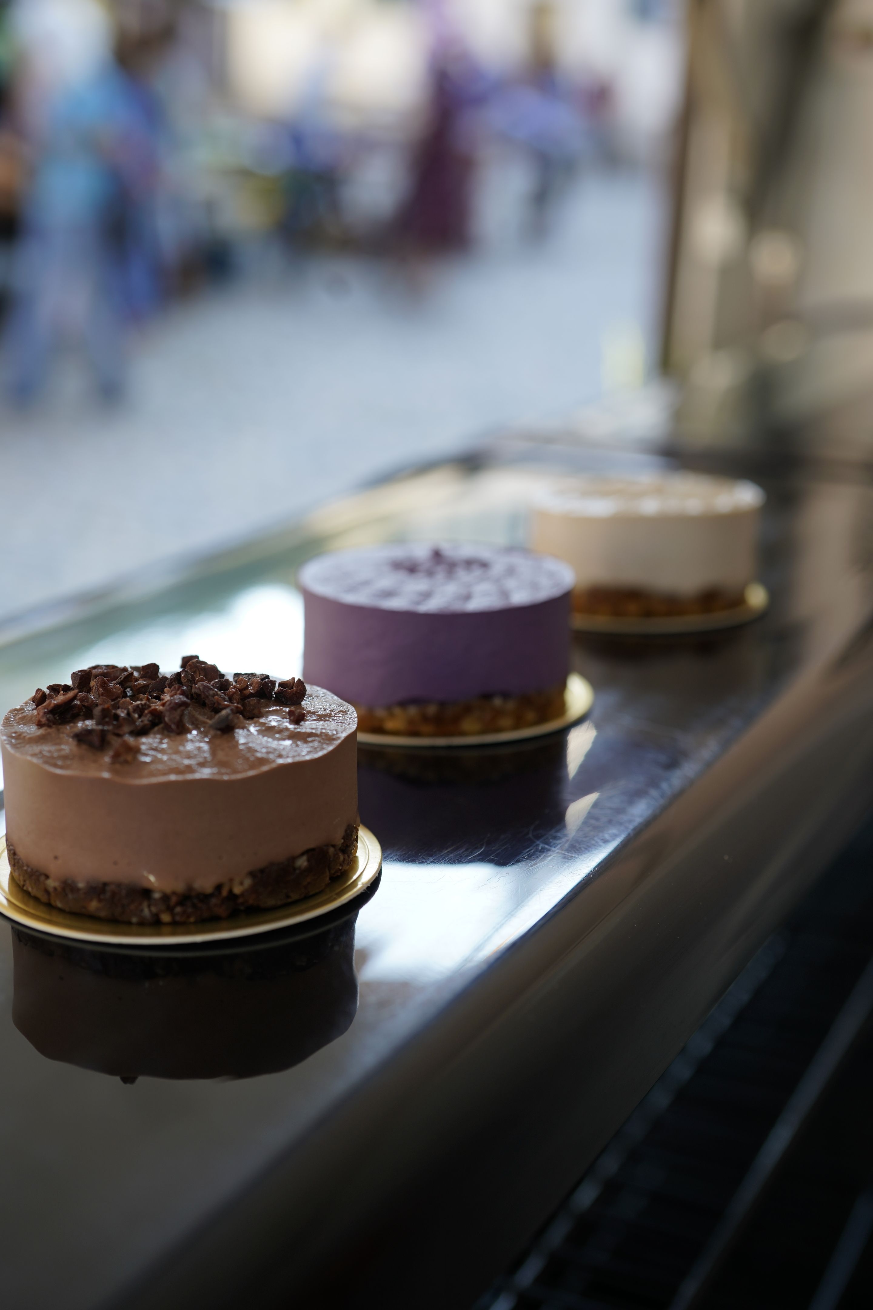 Drei kleine Torten in verschiedenen Farben stehen auf einem glänzenden Tablett. Der Hintergrund ist unscharf und zeigt eine belebte Straße.
