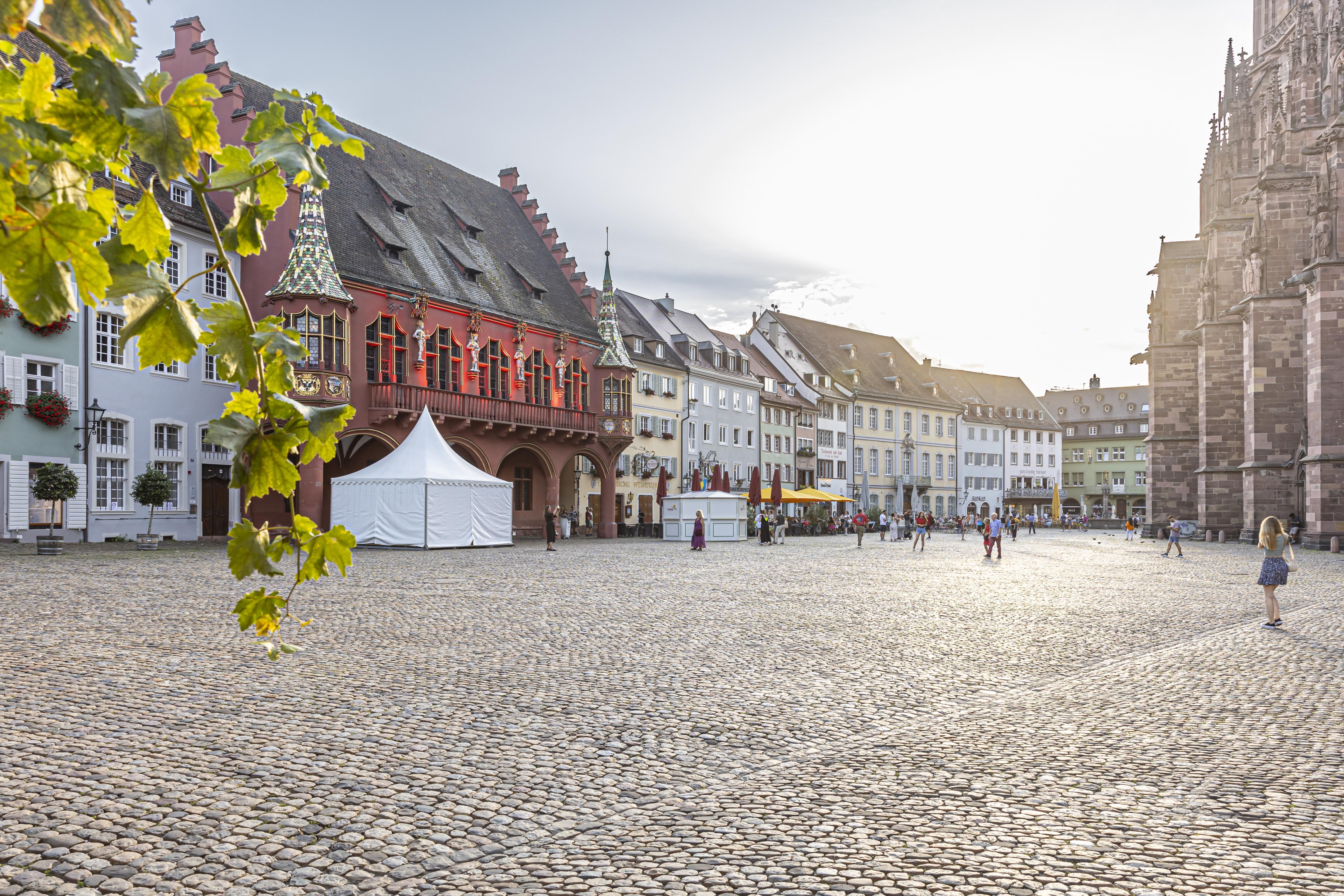 Das Historische Kaufhaus am Münsterplatz in Freiburg bei Sonnenschein. Menschen spazieren über das Kopfsteinpflaster, im Hintergrund historische Gebäude.