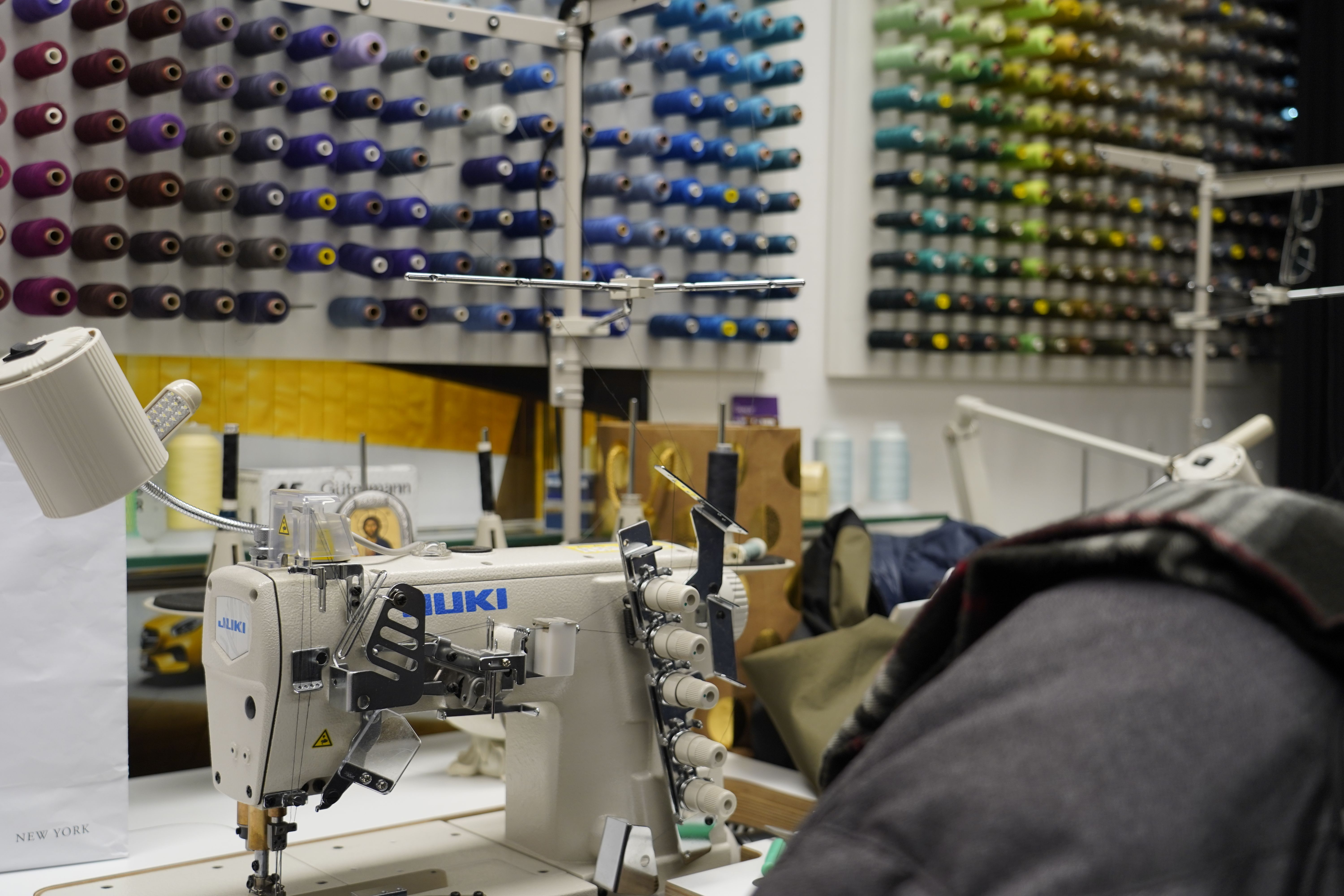 Nähmaschine in einer Schneiderei, umgeben von bunten Garnrollen an der Wand. Ein Arbeitsplatz mit verschiedenen Werkzeugen und Materialien.