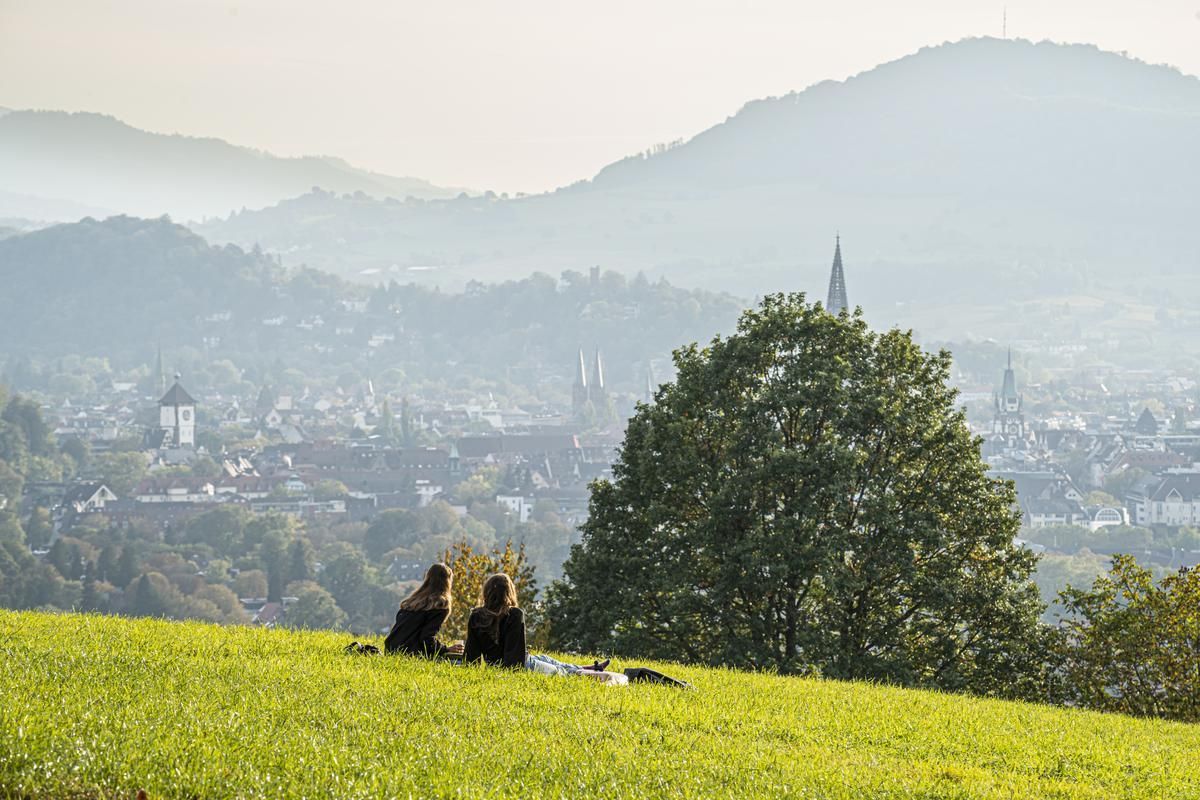 Zwei Personen genießen den Ausblick von einer Wiese auf die Stadt Freiburg im Breisgau, umgeben von grünen Hügeln und Bäumen.