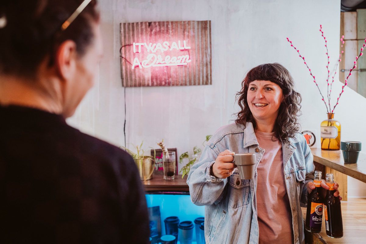 Eine Frau in einer Jeansjacke lächelt und hält eine Tasse und zwei Flaschen in einem Café. Im Hintergrund leuchtet ein Neonschild.