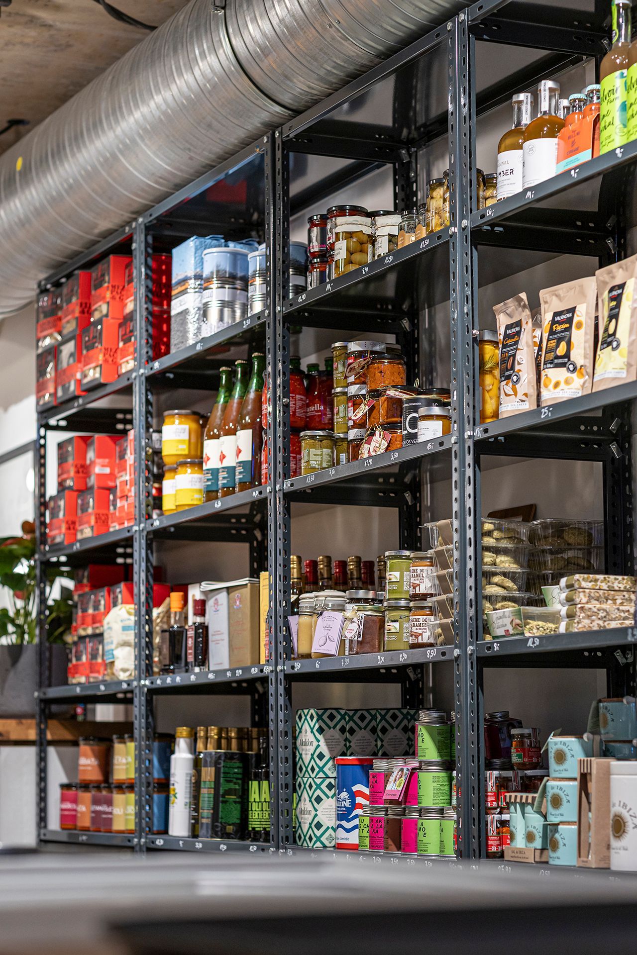 Regale in einem Geschäft, gefüllt mit verschiedenen Lebensmitteln wie Konserven, Flaschen und Verpackungen. Ein Lüftungsrohr verläuft oberhalb der Regale.