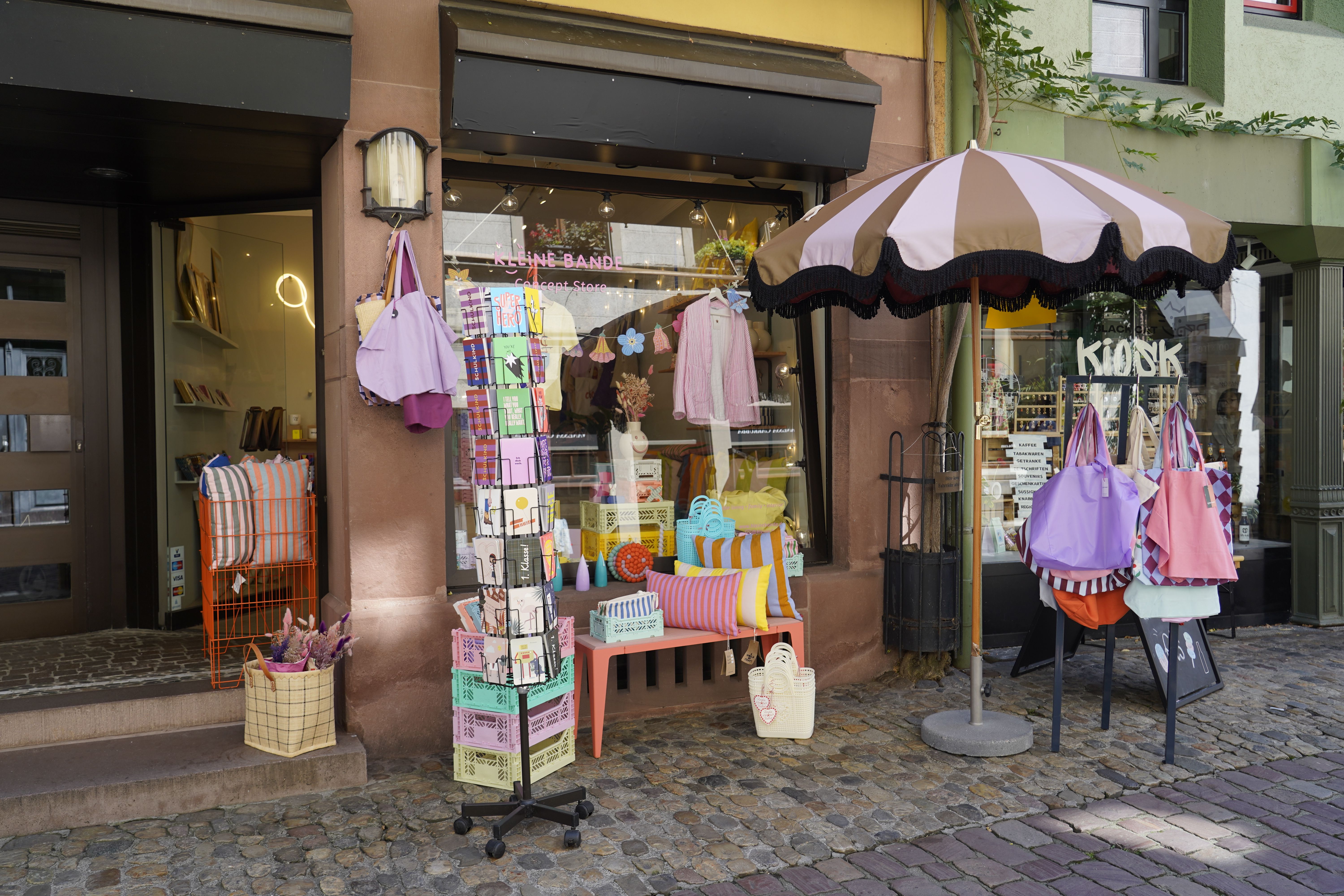 Ein kleiner Laden mit bunten Taschen, Kissen und Kartenständer vor der Tür. Ein gestreifter Sonnenschirm spendet Schatten auf dem Kopfsteinpflaster.