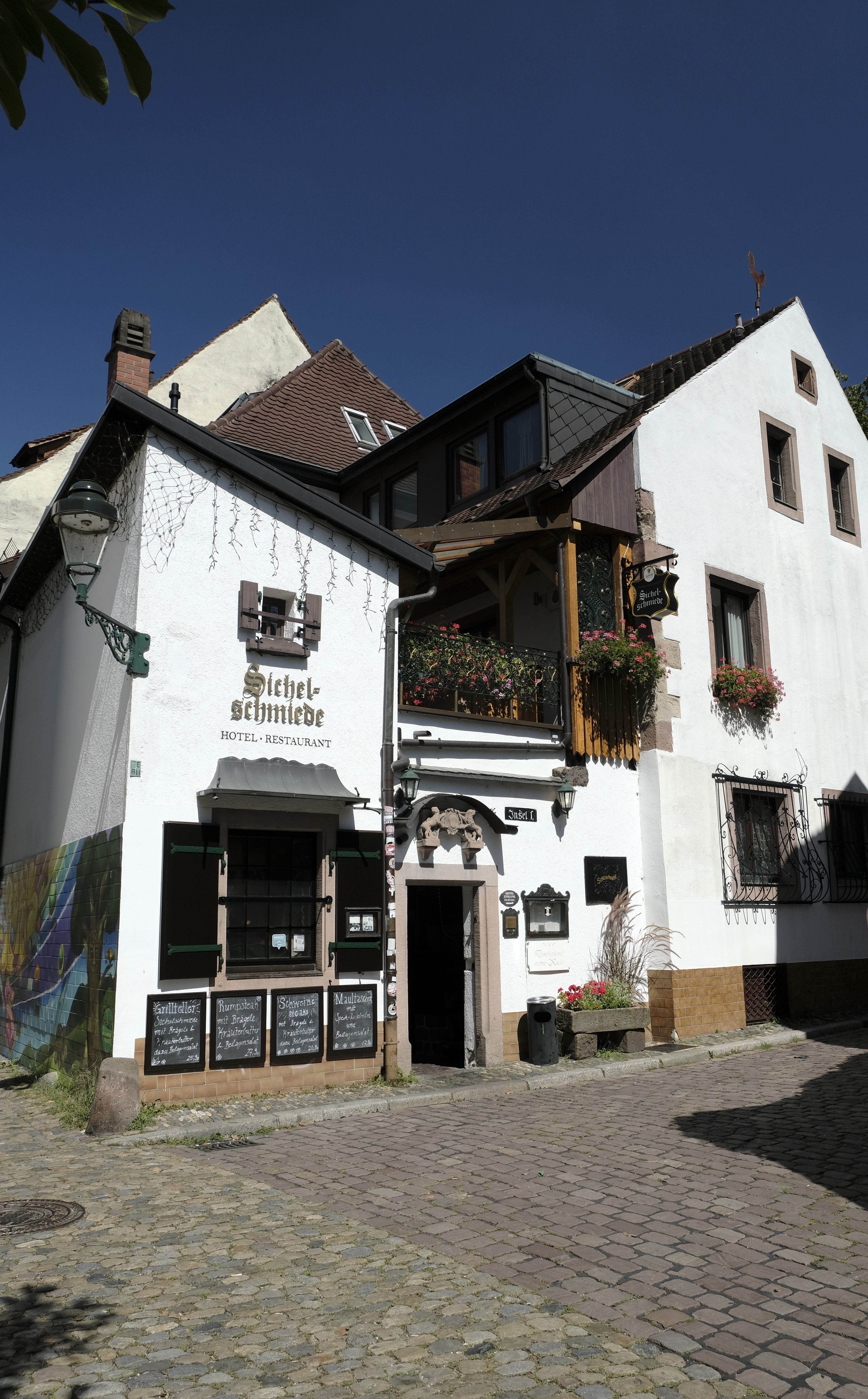 Historisches Gebäude mit der Aufschrift 'Sichelschmiede', Hotel und Restaurant, auf Kopfsteinpflasterstraße bei klarem Himmel.