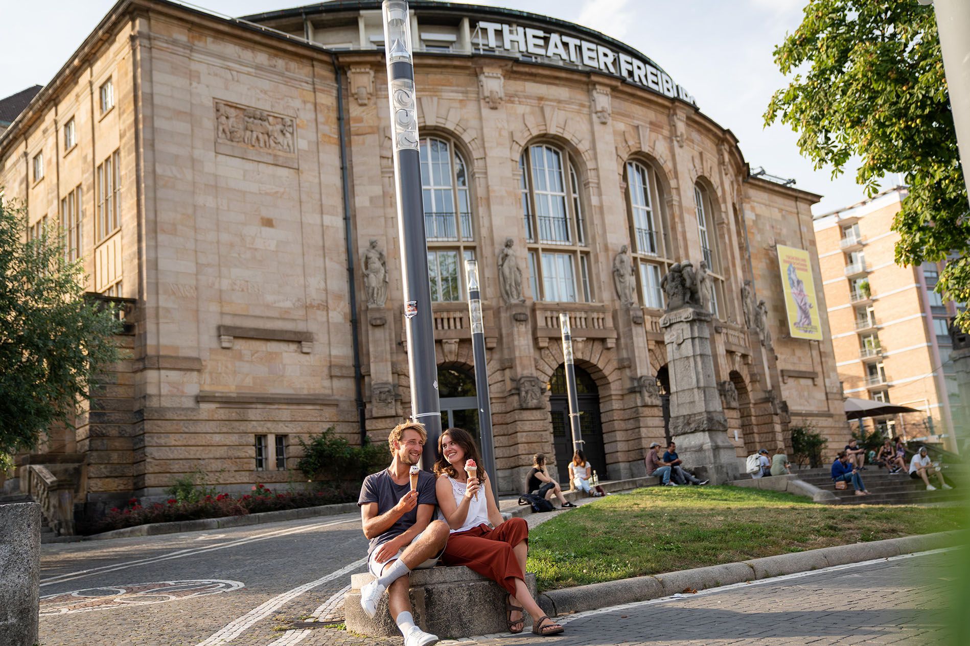 Blick auf das Theater Freiburg, im Vordergrund sitzt ein paar und isst Eis