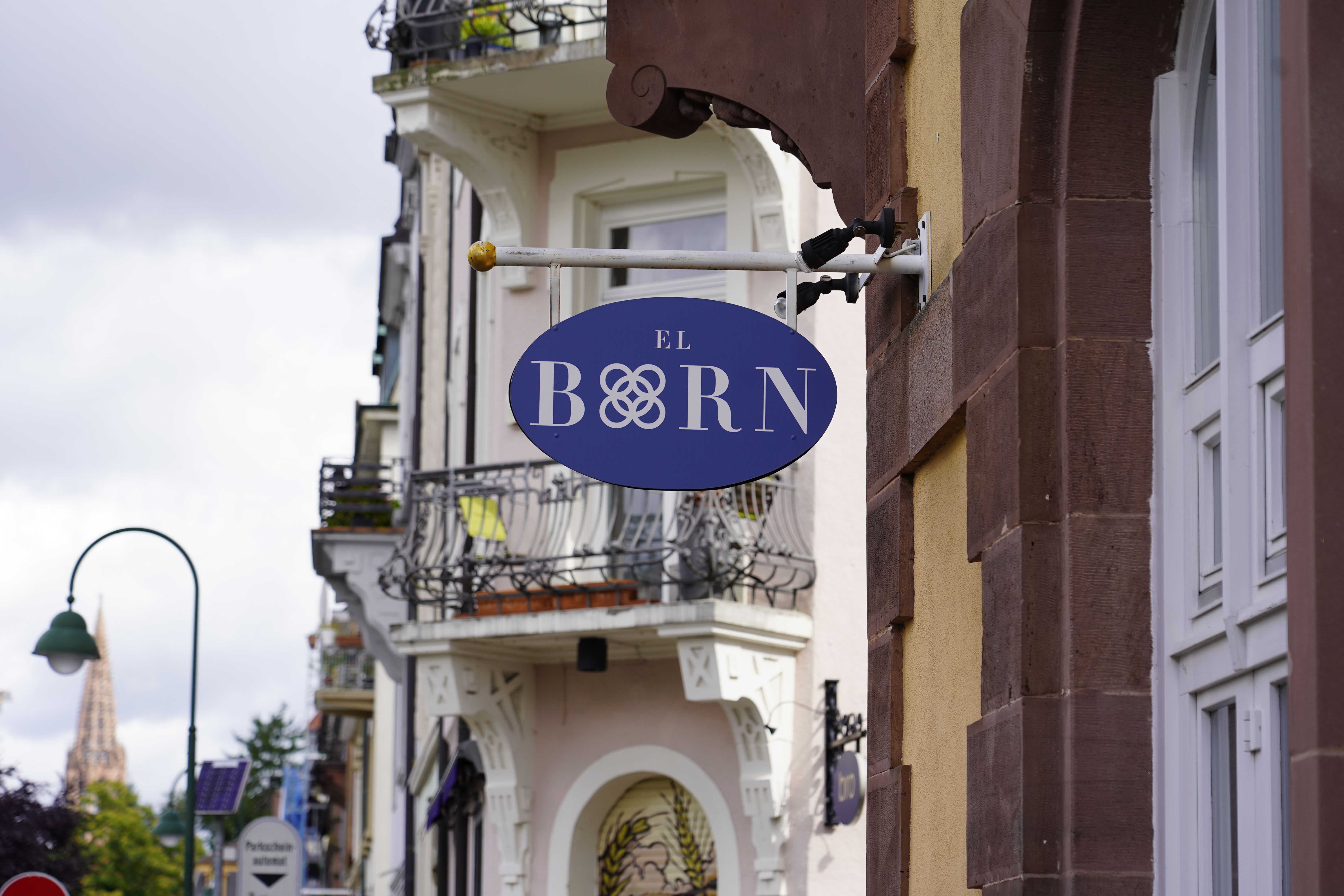 Ein blaues Schild mit der Aufschrift 'El Born' hängt an einer Hauswand in Freiburg. Im Hintergrund sind Balkone und ein Kirchturm sichtbar.
