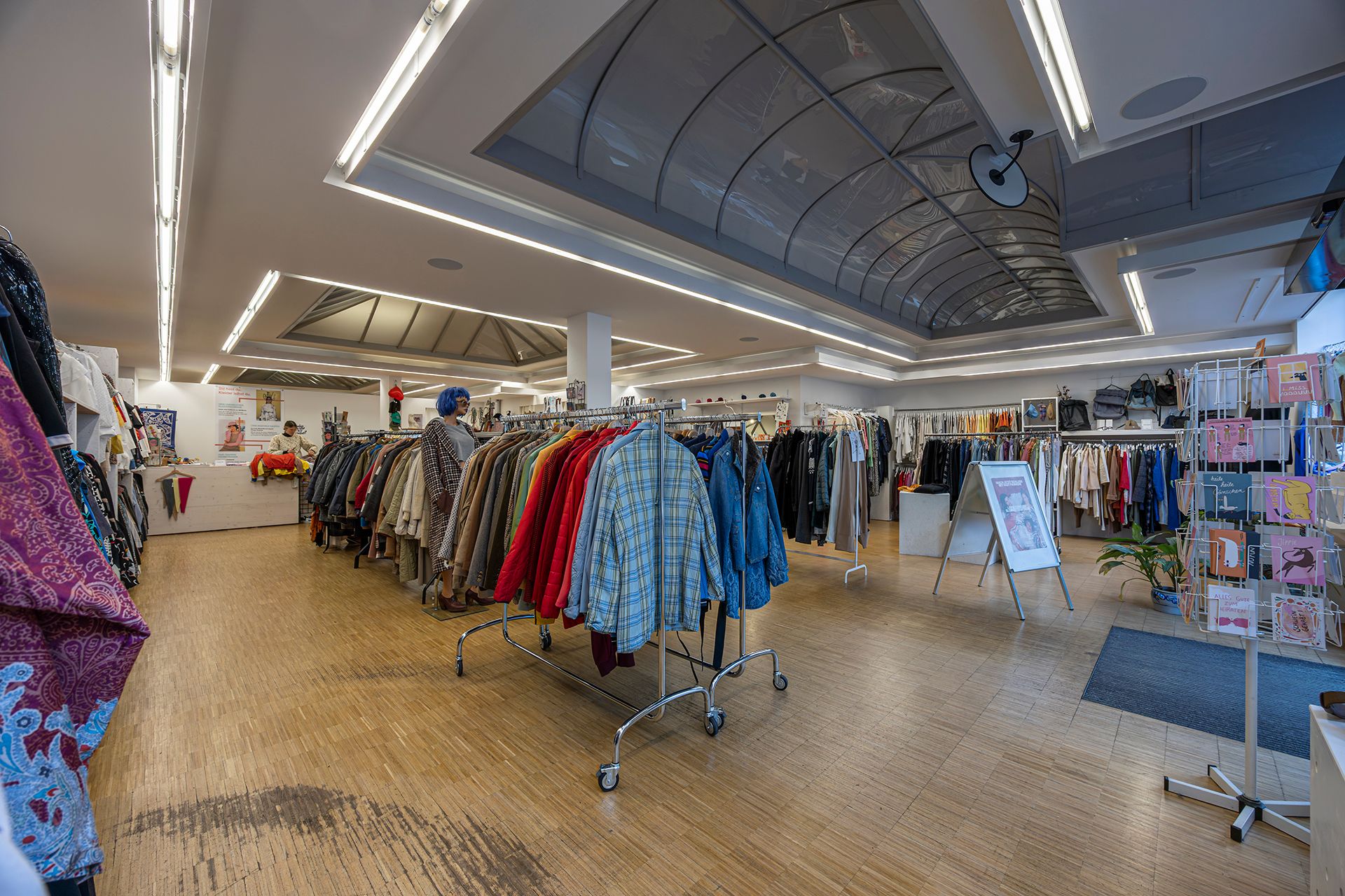 Ein Second-Hand-Laden mit Kleiderständern voller bunter Kleidung. Eine Schaufensterpuppe trägt eine blaue Perücke. Helle Beleuchtung und Holzboden.