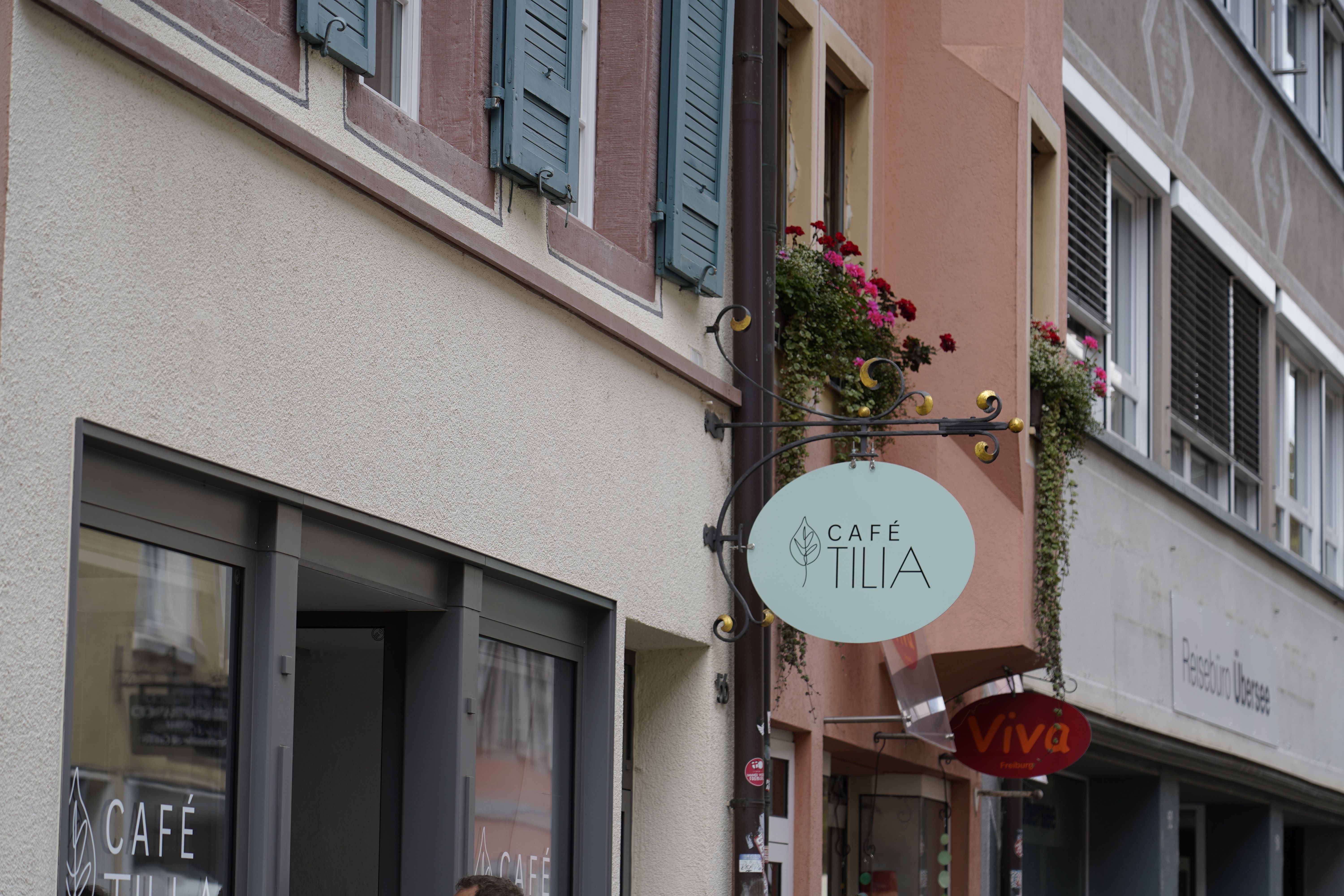Außenansicht des Café Tilia in Freiburg. Ein ovales Schild hängt an der Fassade, daneben Fenster mit Blumen. 