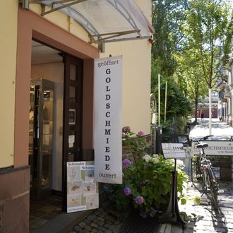 Eingang einer Goldschmiede mit Schild 'geöffnet'. Vor dem Laden stehen Fahrräder und Pflanzen. Die Straße ist gepflastert und von Bäumen gesäumt.