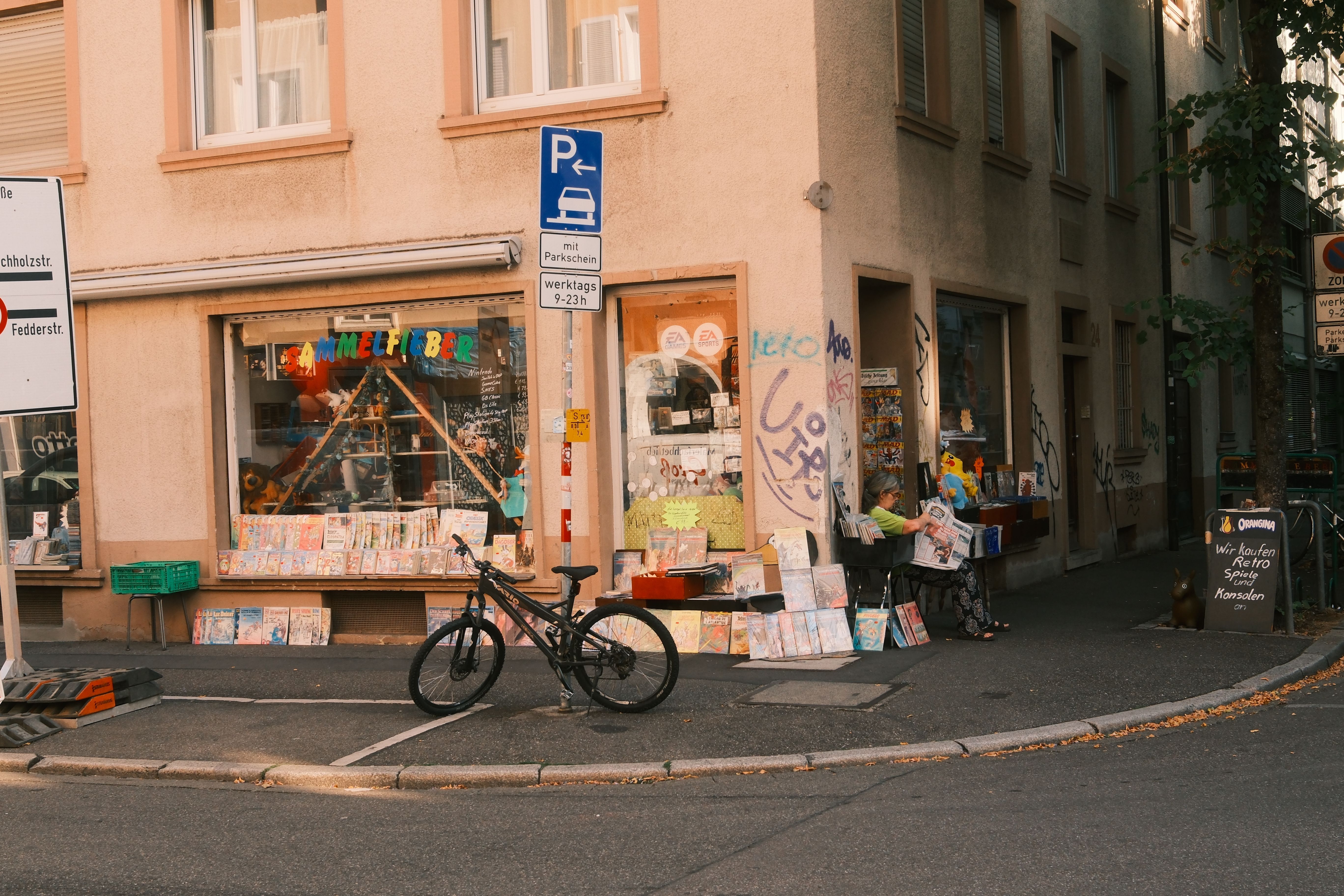 Straßenecke mit dem Laden „Sammelfieber“, ausliegenden Comics und Spielen, Fahrrad davor und einer sitzenden Person am Eingang.