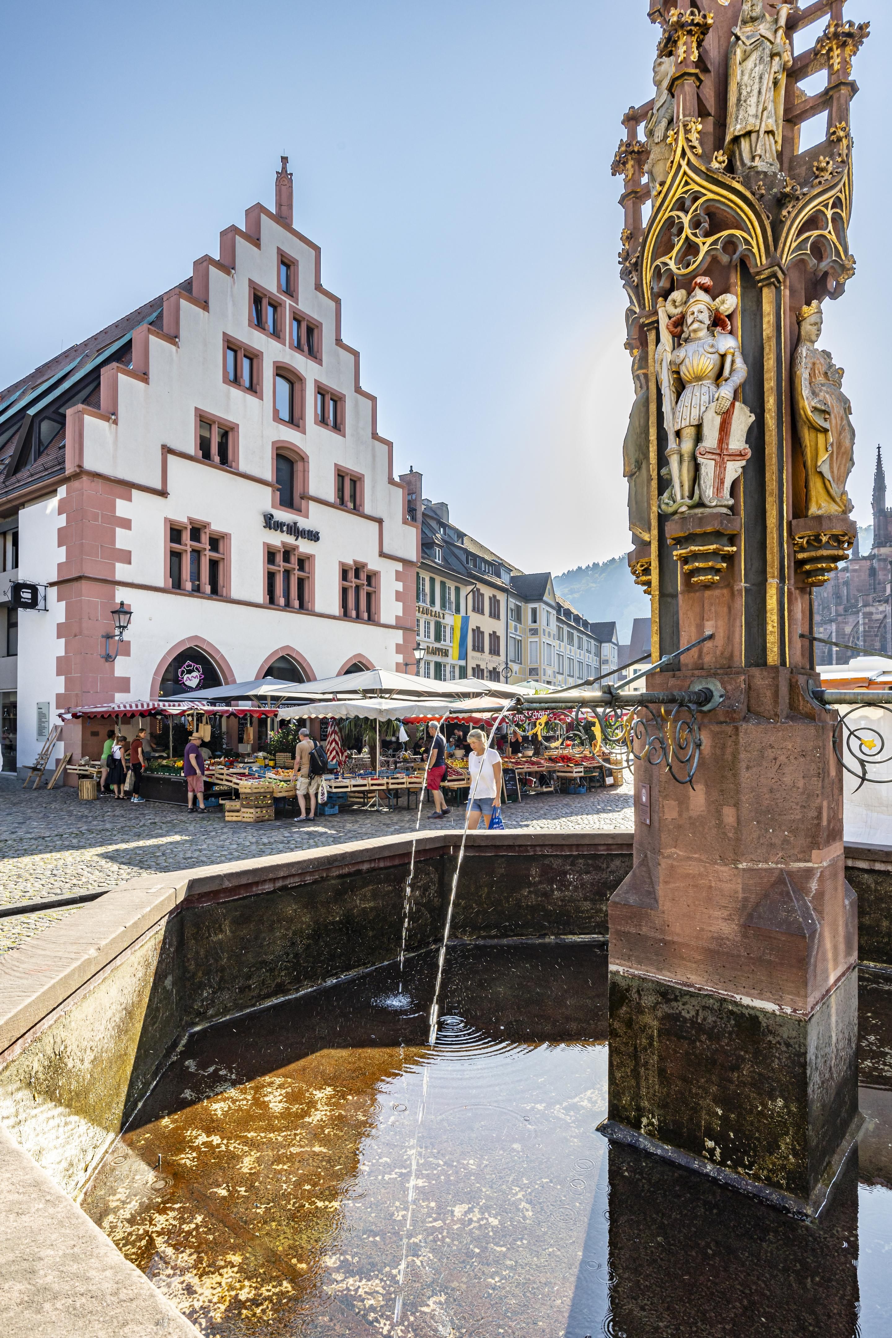 Ein Brunnen mit kunstvollen Figuren im Vordergrund, dahinter ein historisches Gebäude mit Treppengiebel und ein Markt mit Ständen und Menschen.