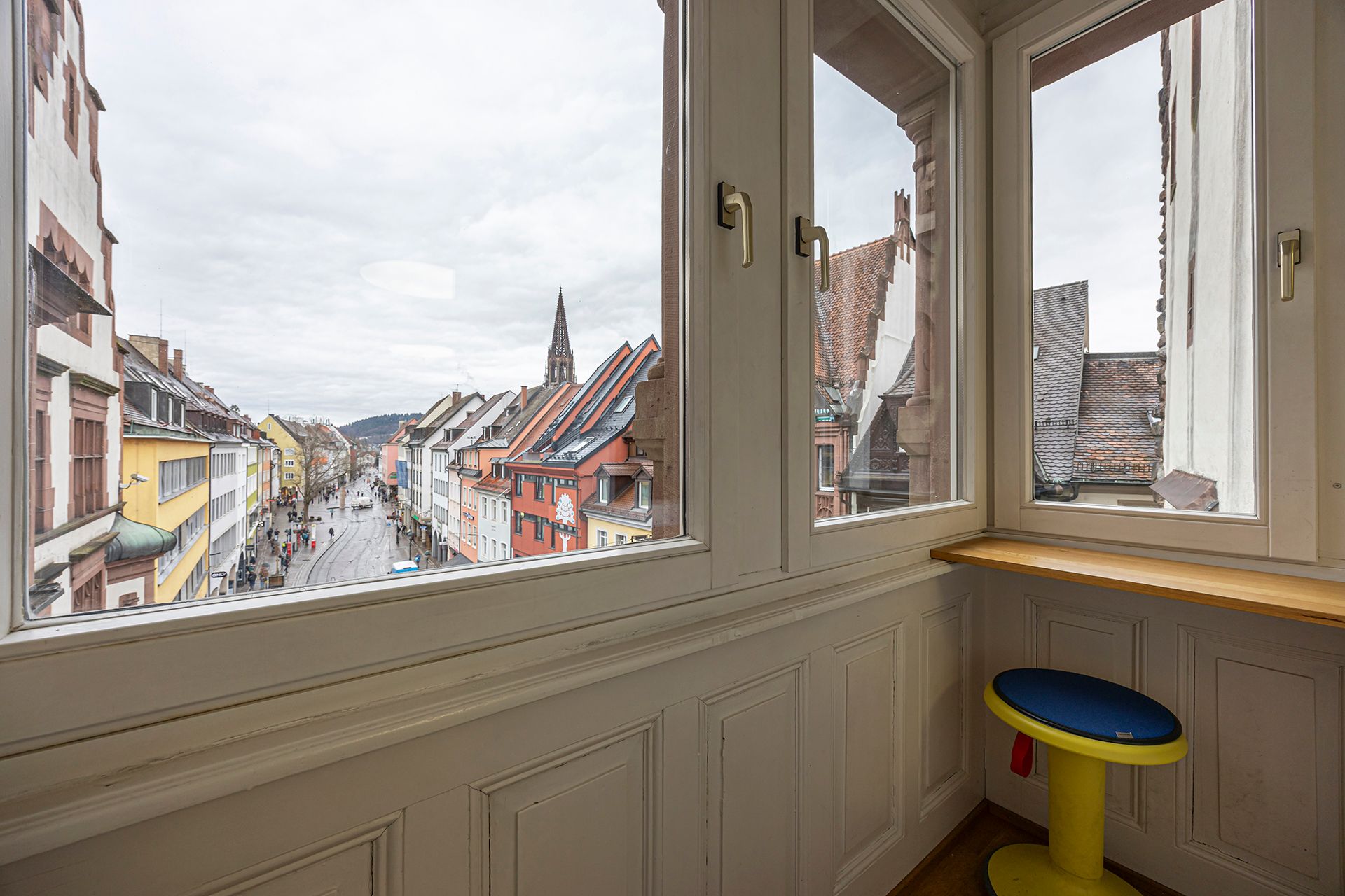 Ausblick aus einem Fenster auf eine belebte Straße in Freiburg. Bunte Häuser und ein Kirchturm sind zu sehen. Ein gelber Hocker steht am Fenster.