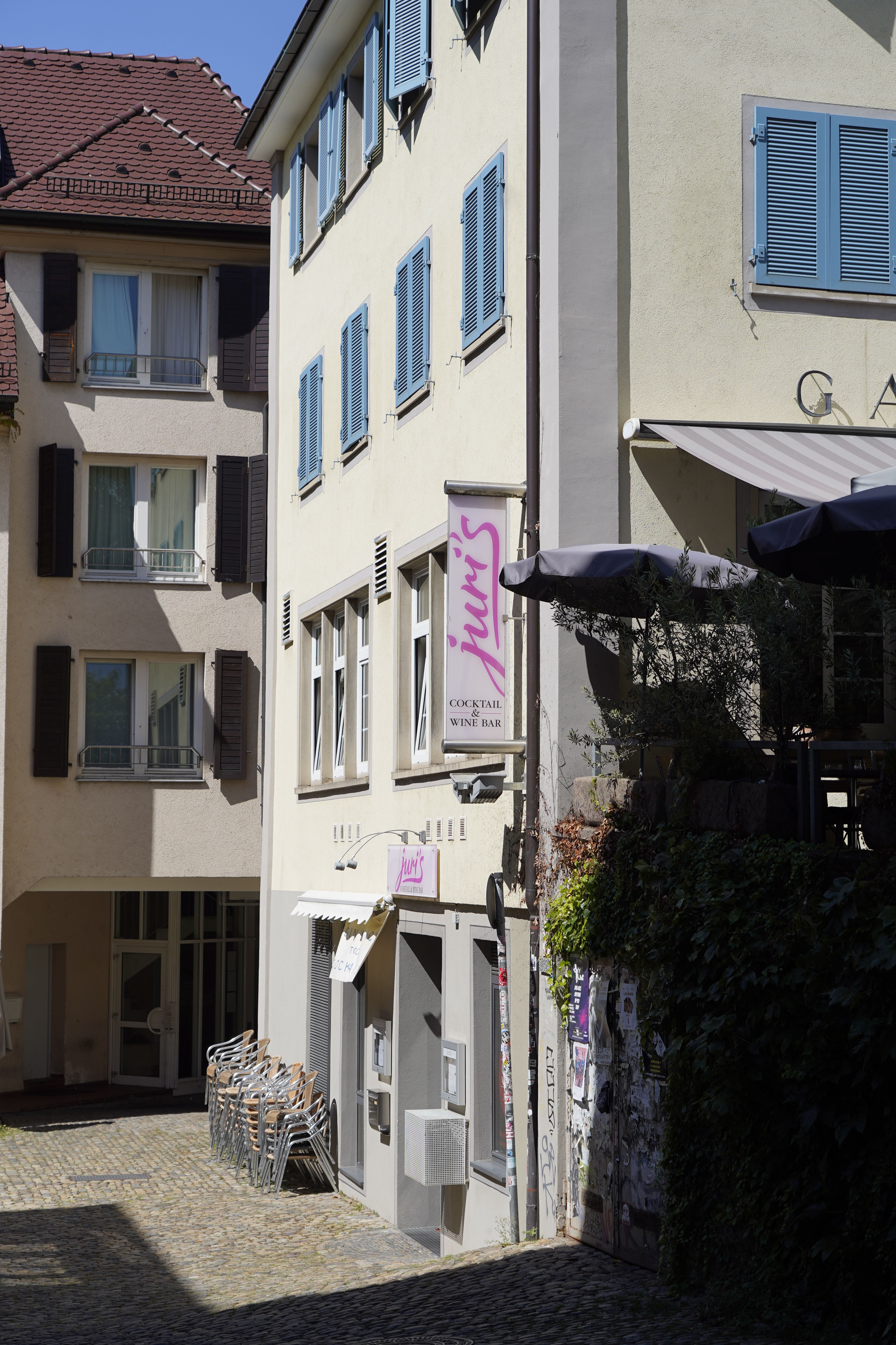 Außenansicht einer Cocktail- und Weinbar in einer engen Gasse mit gestapelten Stühlen und blauen Fensterläden.