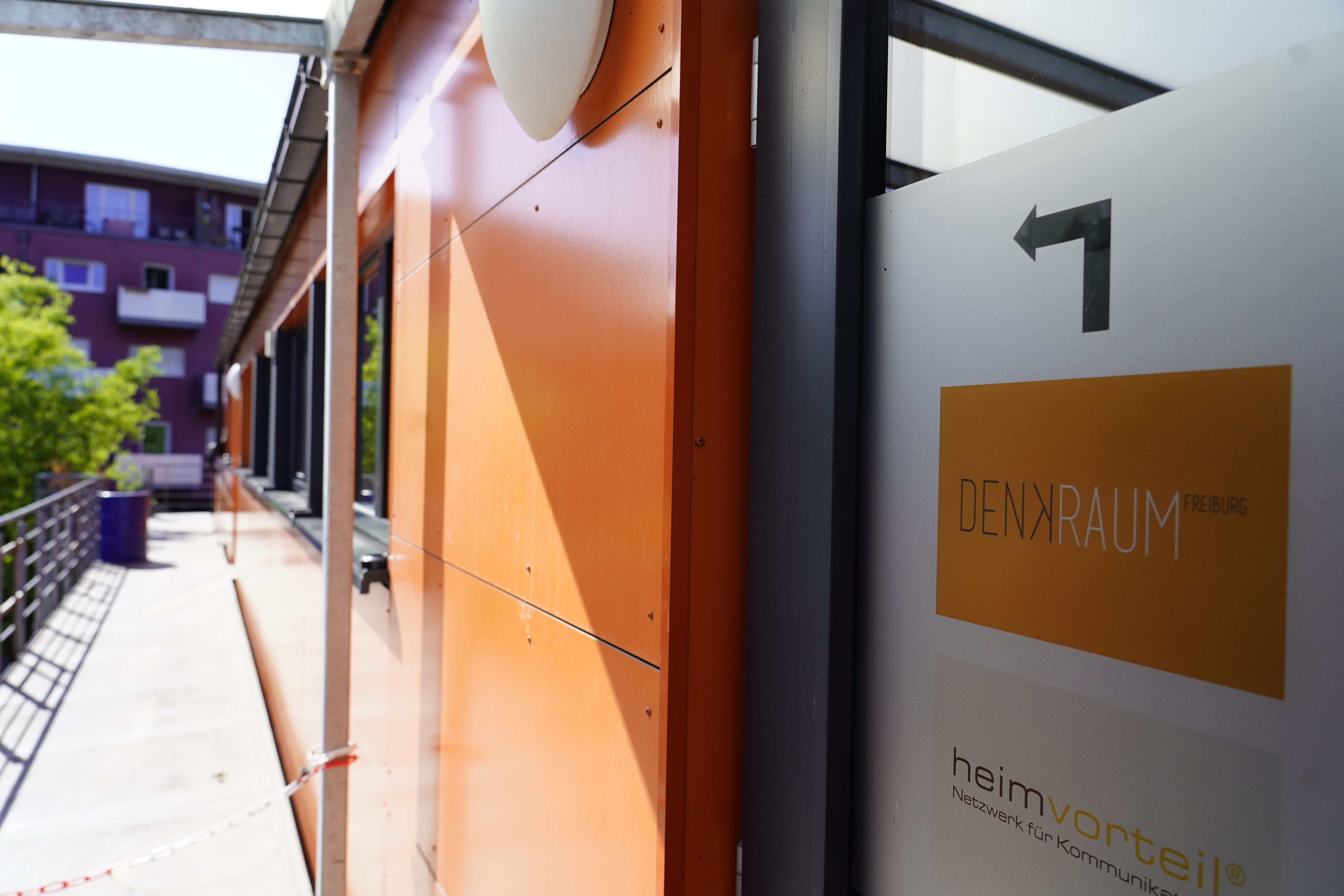 Ein Balkon mit orangefarbener Wand, einem Schild mit der Aufschrift 'DENKRAUM Freiburg' und einem Pfeil. Im Hintergrund sind Gebäude und Bäume sichtbar.