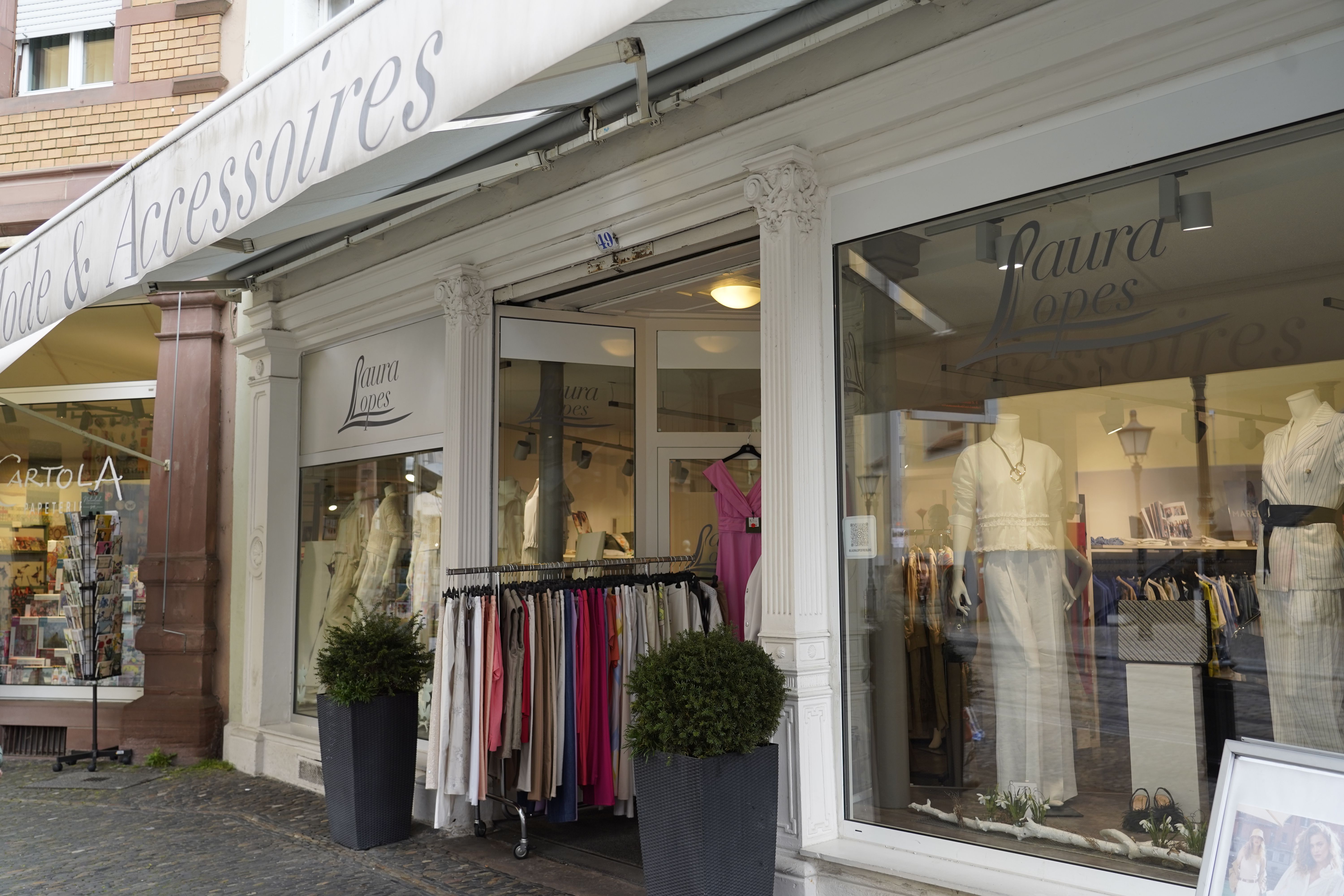 Modegeschäft 'Laura Lopes' mit Schaufenster, Kleiderständer und Pflanzen vor dem Eingang. Schaufenster zeigt Mannequins in eleganter Kleidung.