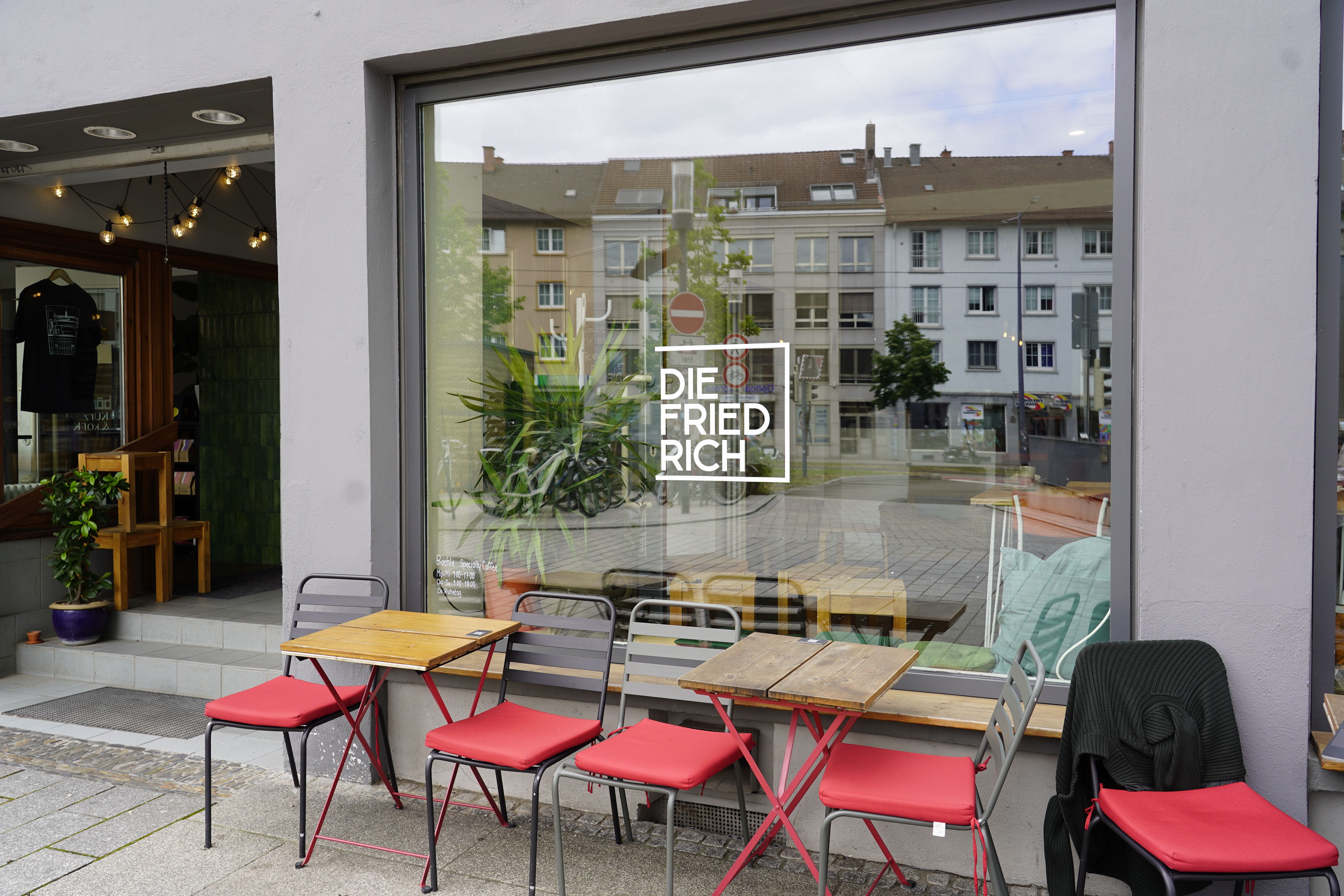 Außenansicht des Do-Workings mit roten Stühlen und Holztischen vor dem Fenster.
