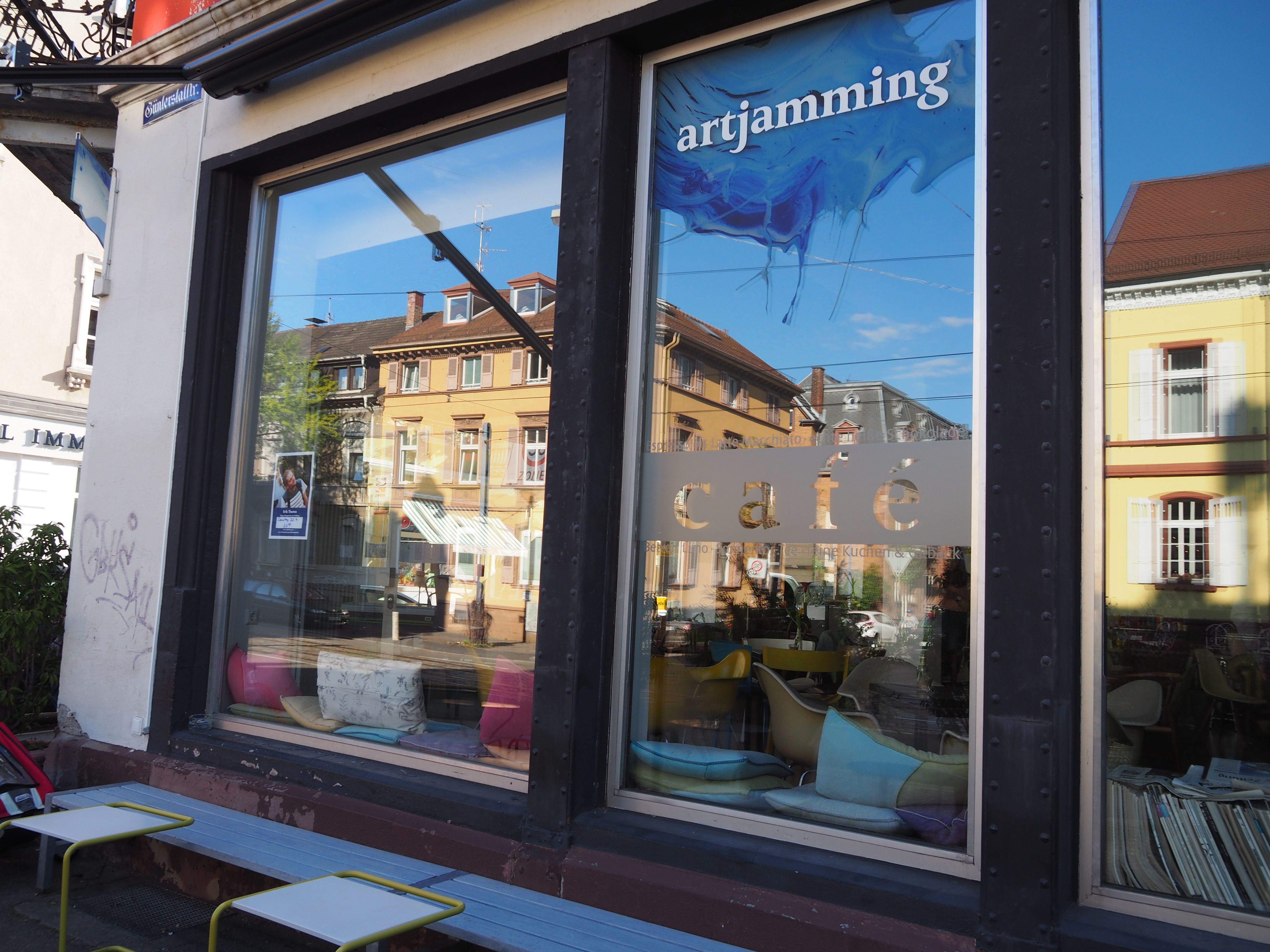 Außenansicht eines Cafés in Freiburg mit großen Fenstern und der Aufschrift 'artjamming'. Innen sind bunte Kissen und Stühle zu sehen.
