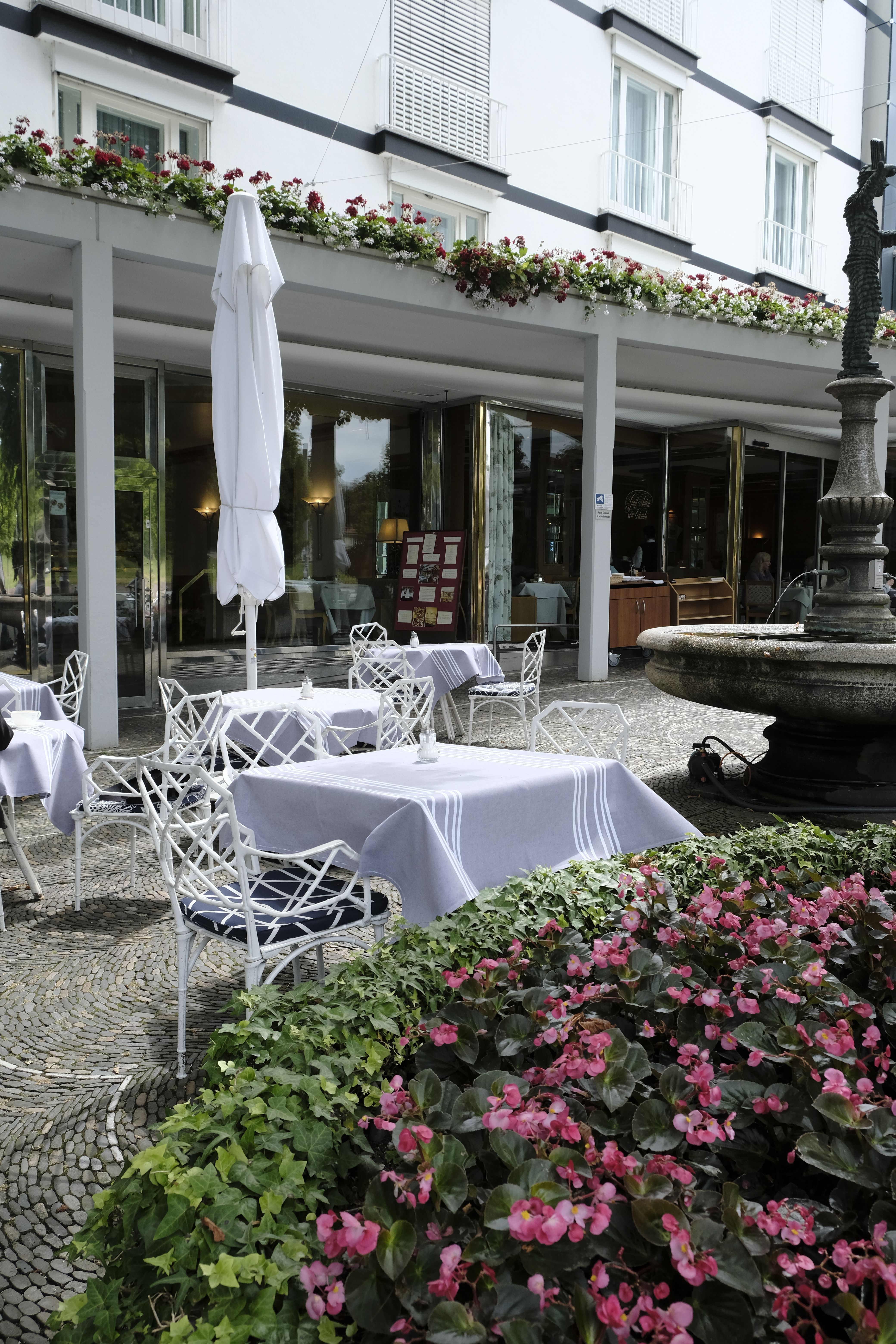 Außenbereich des Cafés mit weißen Tischen und Stühlen, Blumenbeeten und einem Brunnen. Das Gebäude im Hintergrund hat Balkone und Blumenkästen.