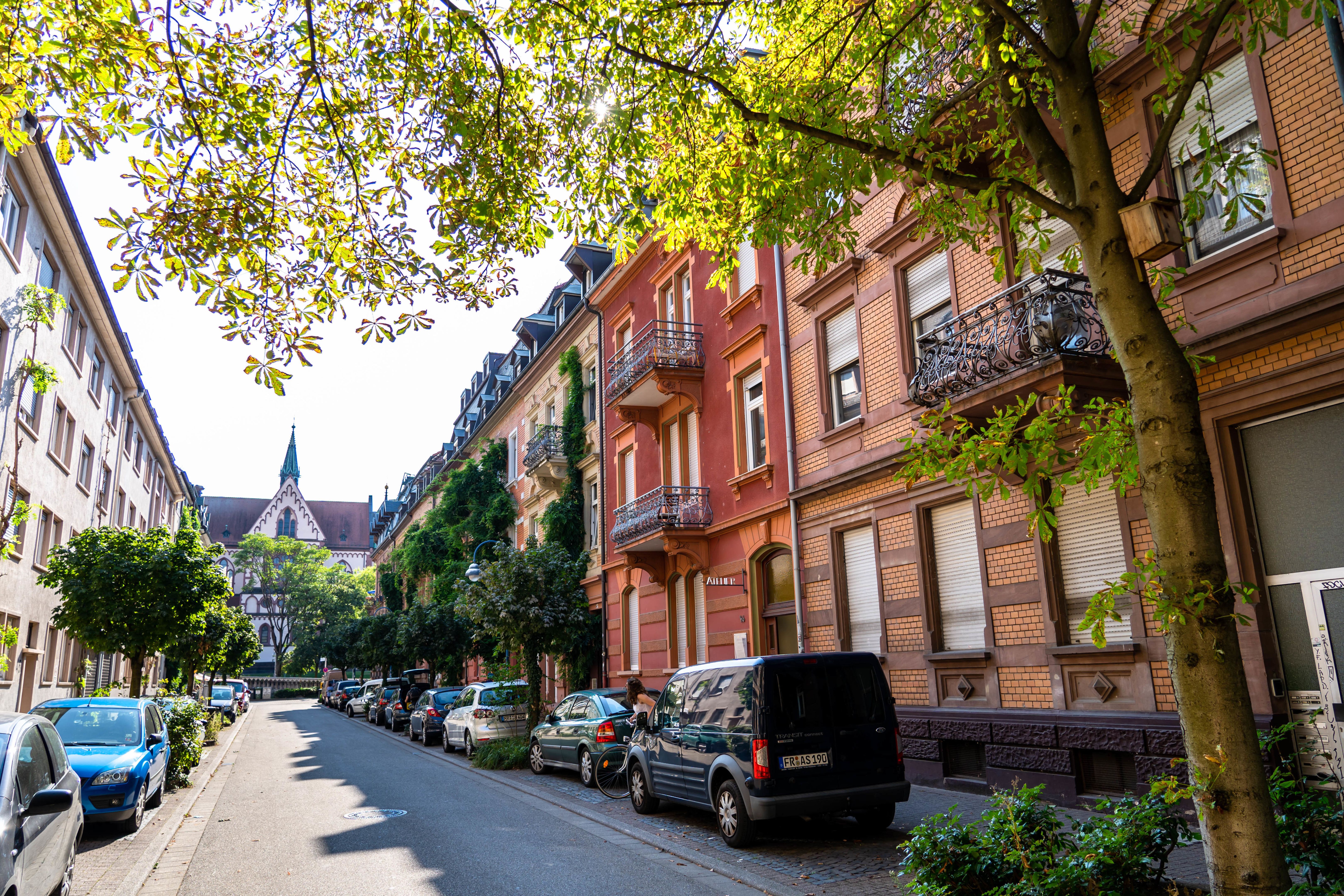 Straße in Freiburg-Stühlinger mit Bäumen, geparkten Autos und einer Kirche im Hintergrund. Die Sonne scheint durch die Blätter der Bäume.