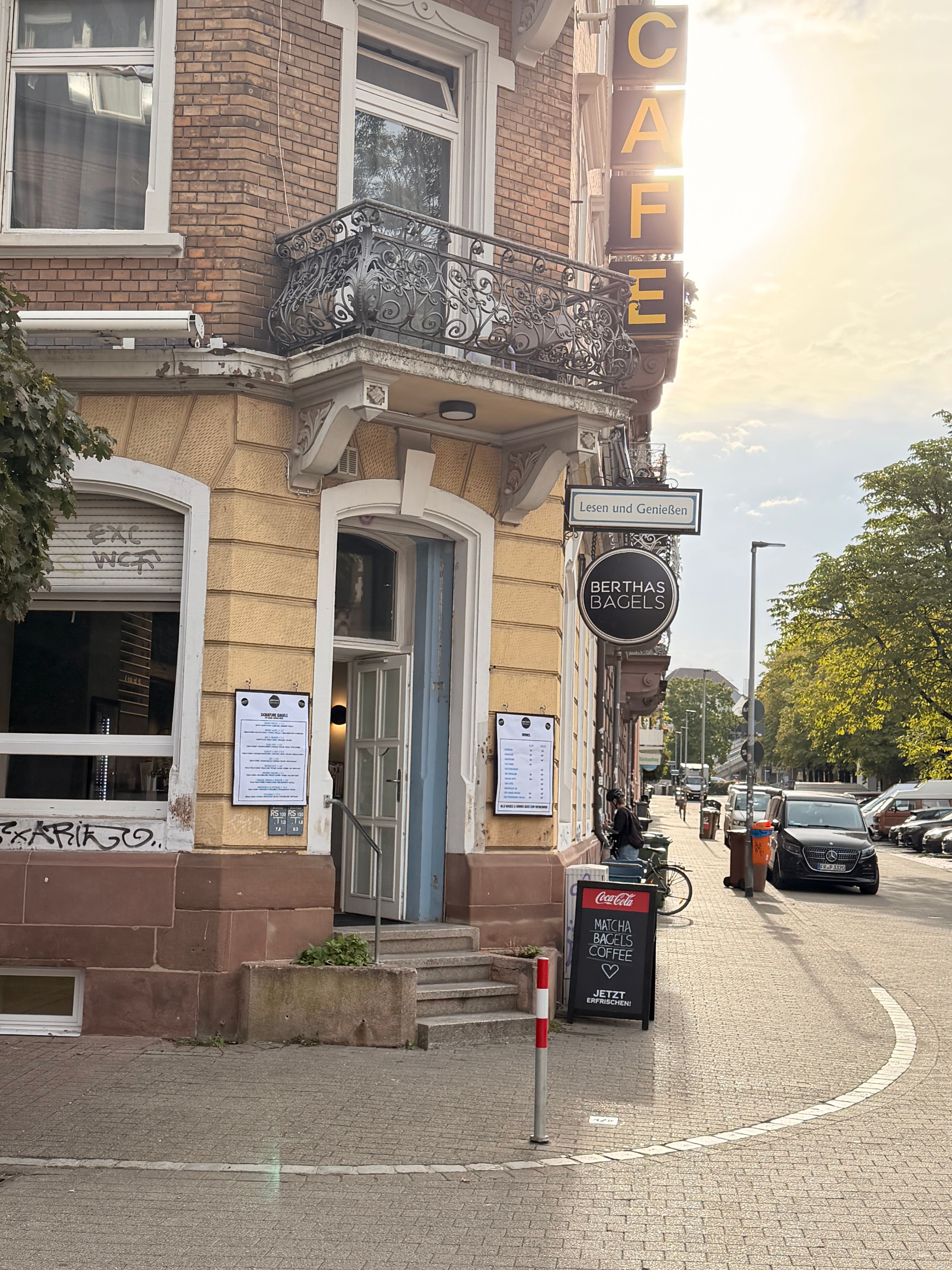 Straßenecke mit Berthas Bagels im Altbau, Café-Schildern und Balkon; warmes Abendlicht an der ruhigen Straße.