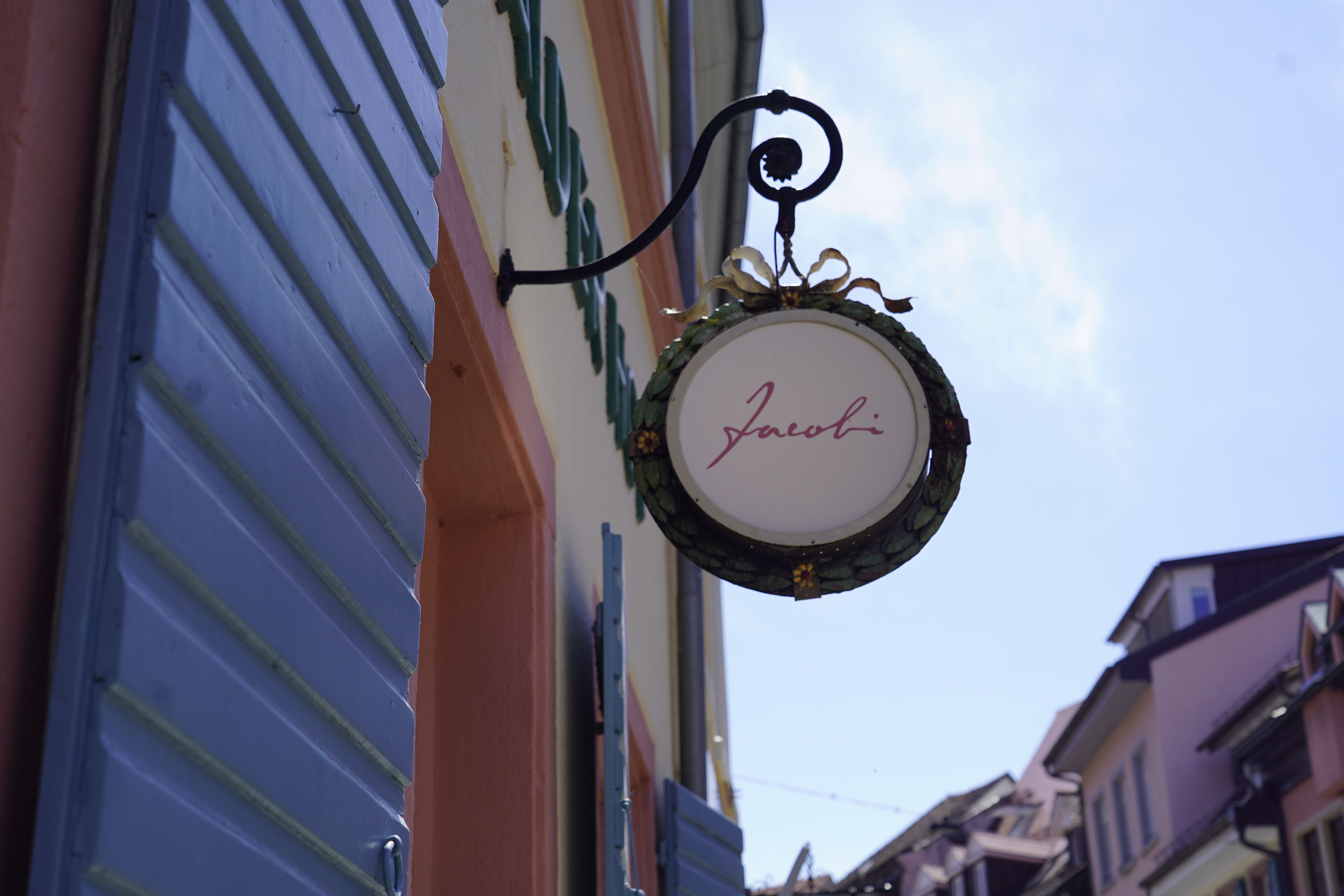 Schild mit 'Jacobi' an einer Hauswand mit blauen Fensterläden, im Hintergrund Dächer und blauer Himmel.