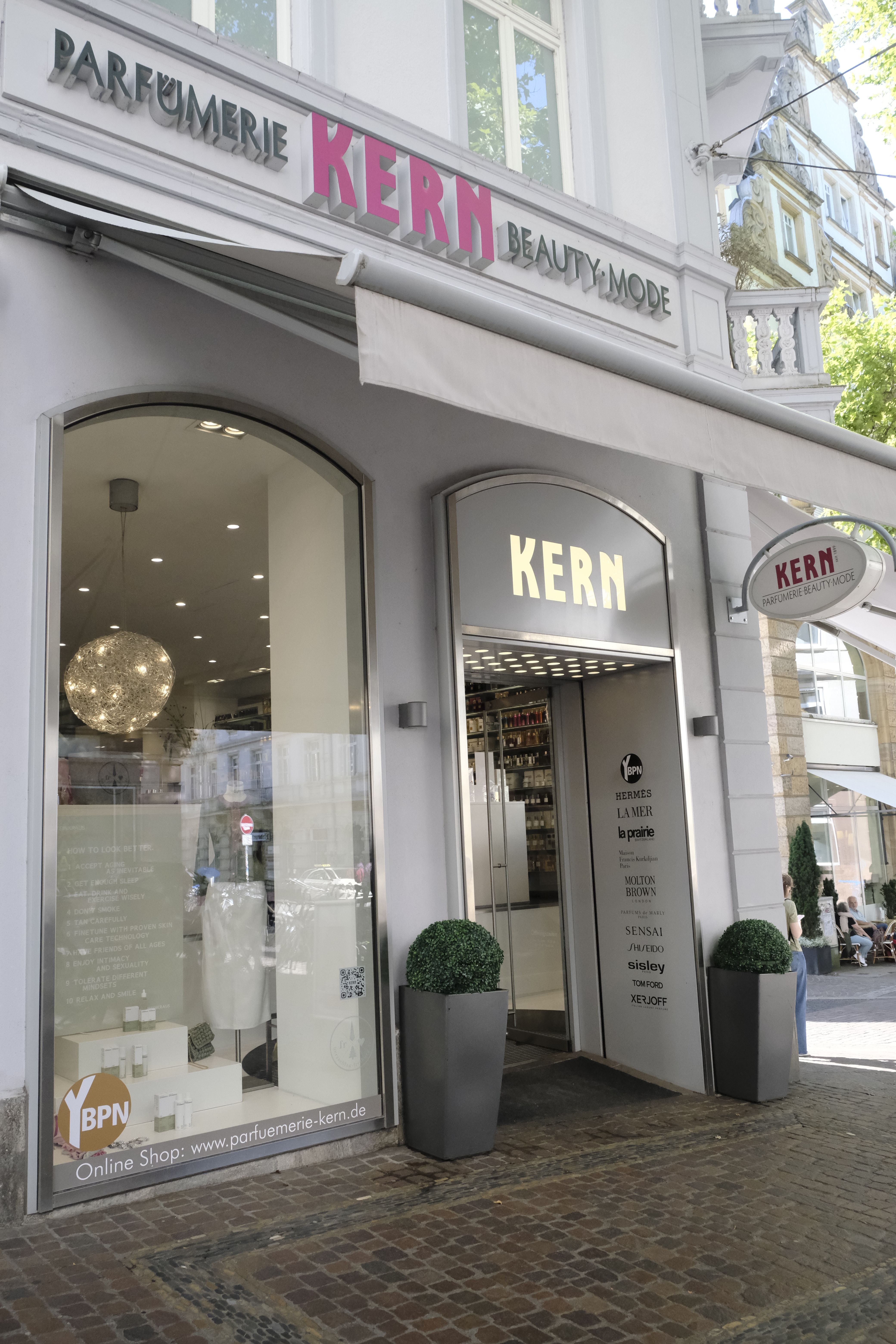 Eingang der Parfümerie Kern mit Schaufenster, Markise und Pflanzendekoration. Das Geschäft bietet Beauty- und Modeprodukte an.