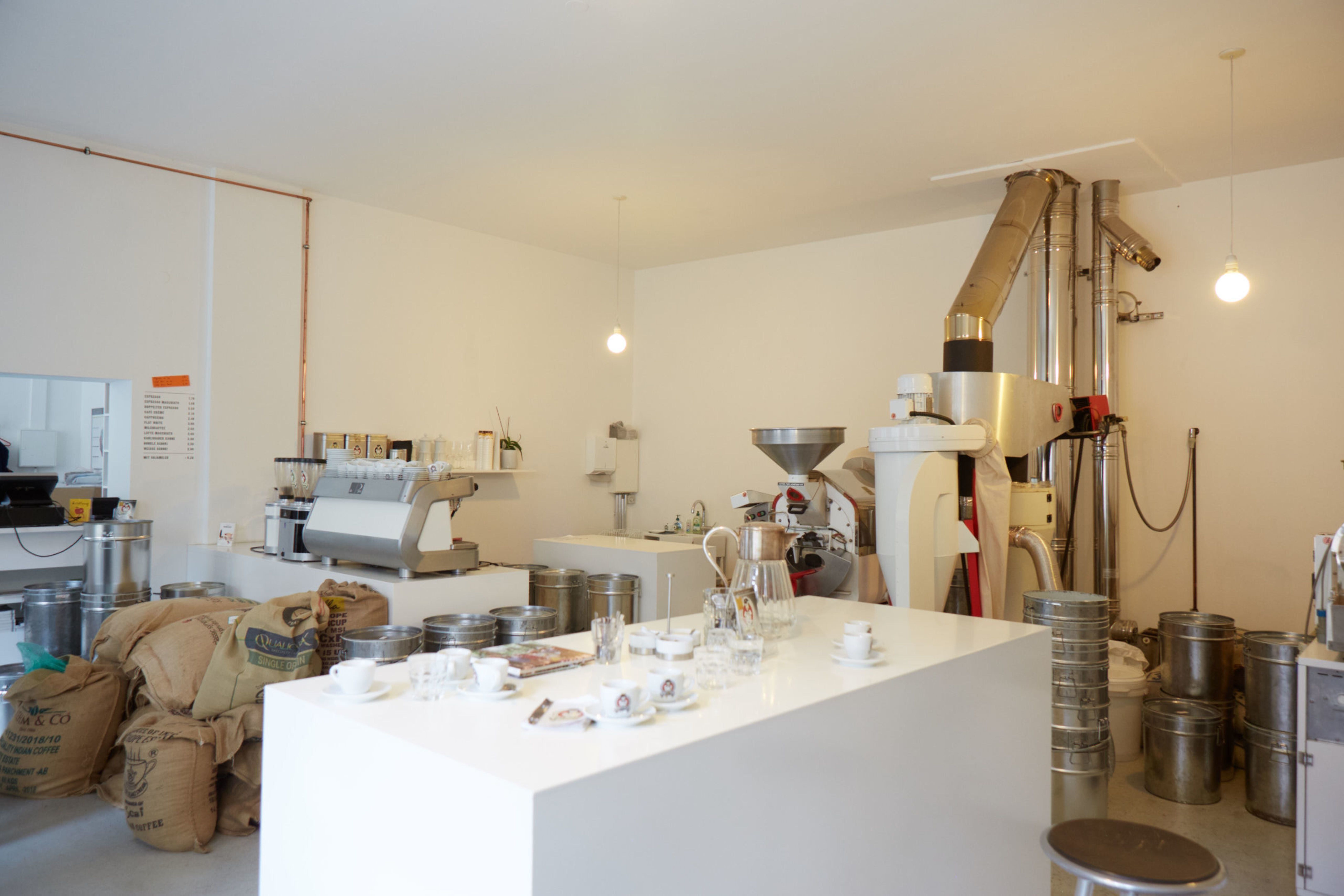 Innenansicht einer Kaffeerösterei mit Röstmaschine, Kaffeesäcken und Tassen auf einem Tisch.