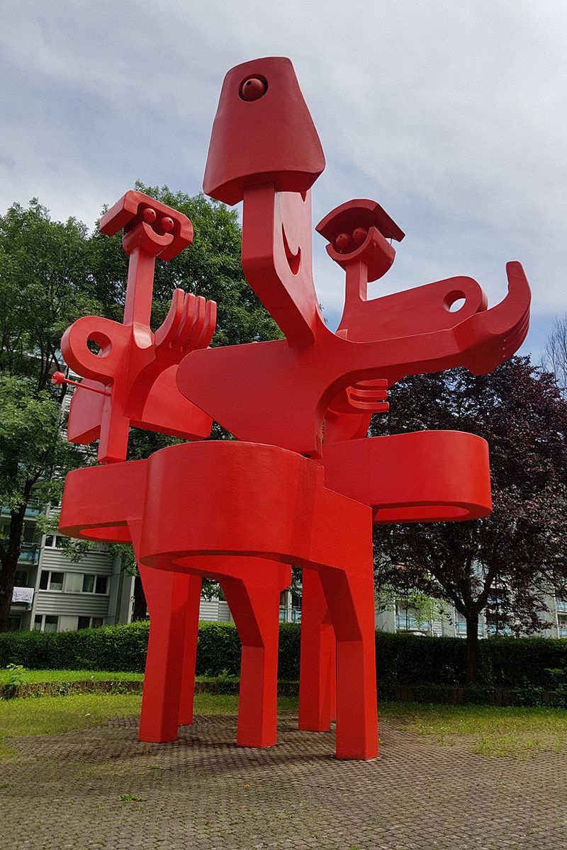 Abstrakte rote Skulptur mit mehreren Armen und Formen, umgeben von Bäumen und Gebäuden im Hintergrund.