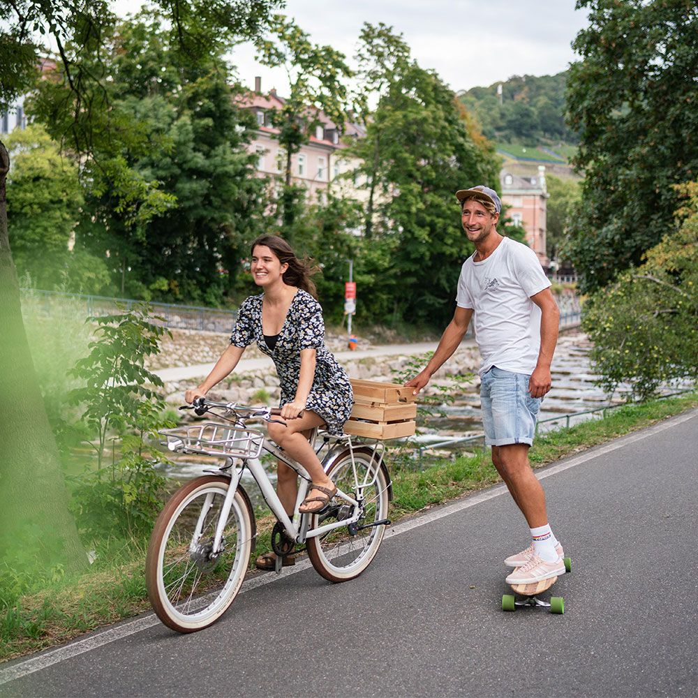Freiburg Dreisamufer Frau im Sommerkleid fährt Fahrrad, Mann fährt auf dem Skateboard nebenher