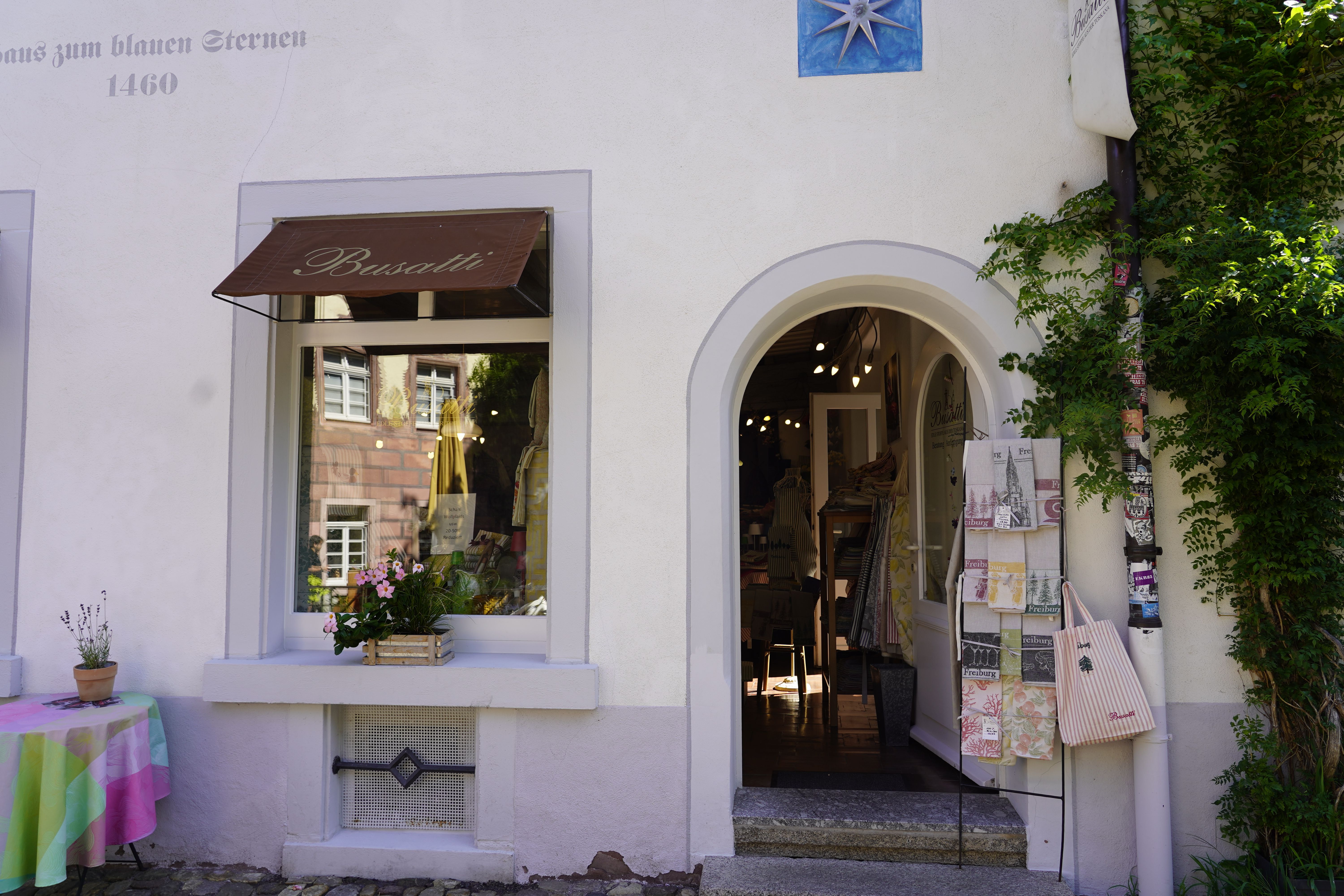 Außenansicht eines Ladens namens Busatti in Freiburg. Ein Schaufenster mit Blumen, ein Eingang mit Textilien und eine mit Pflanzen bewachsene Fassade.