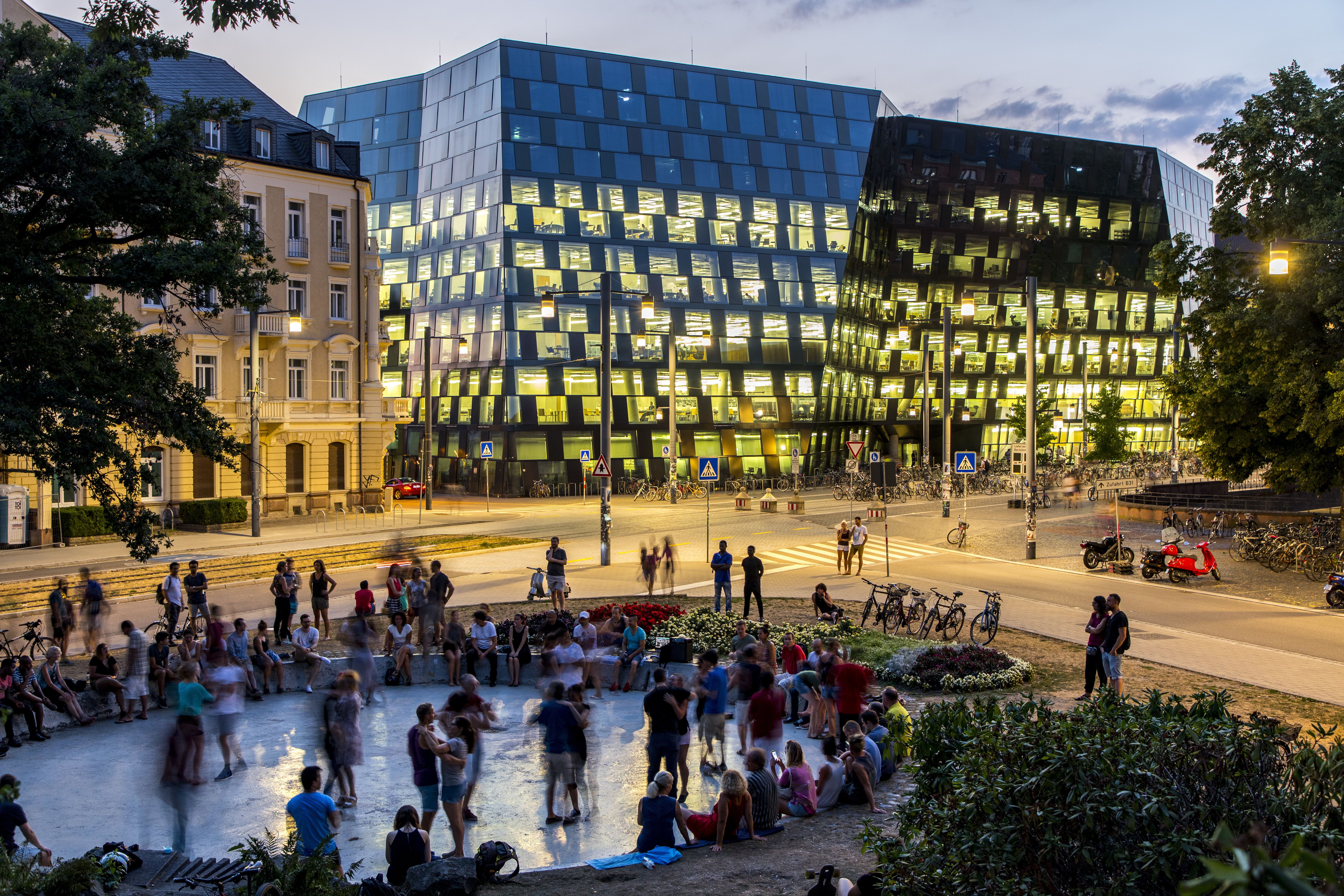 Abendstimmung vor der beleuchteten Universitätsbibliothek Freiburg. Viele Menschen sitzen und stehen im Vordergrund, während die Bibliothek im Hintergrund leuchtet.
