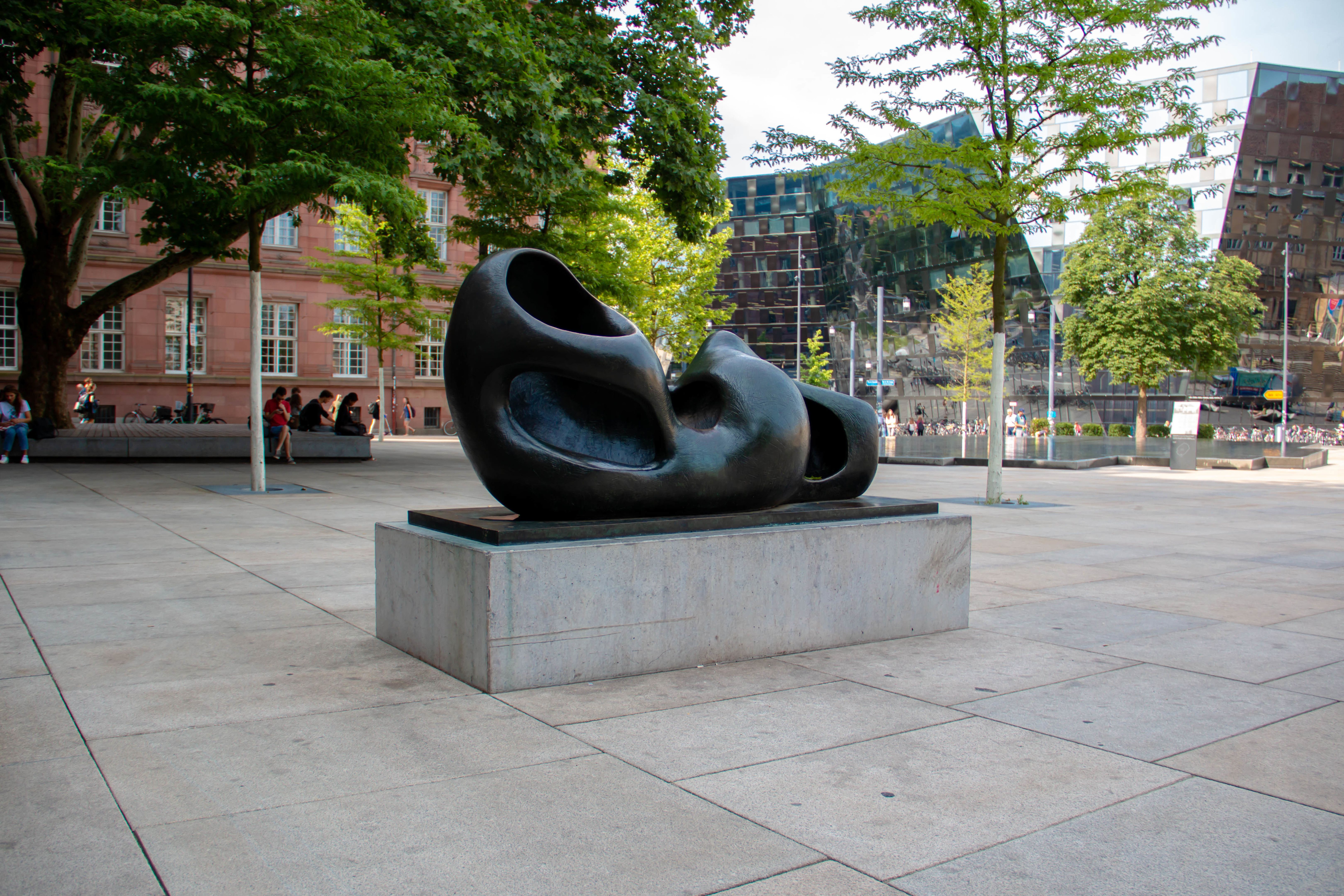 Moderne Skulptur auf einem Platz in Freiburg, umgeben von Bäumen und Gebäuden. 