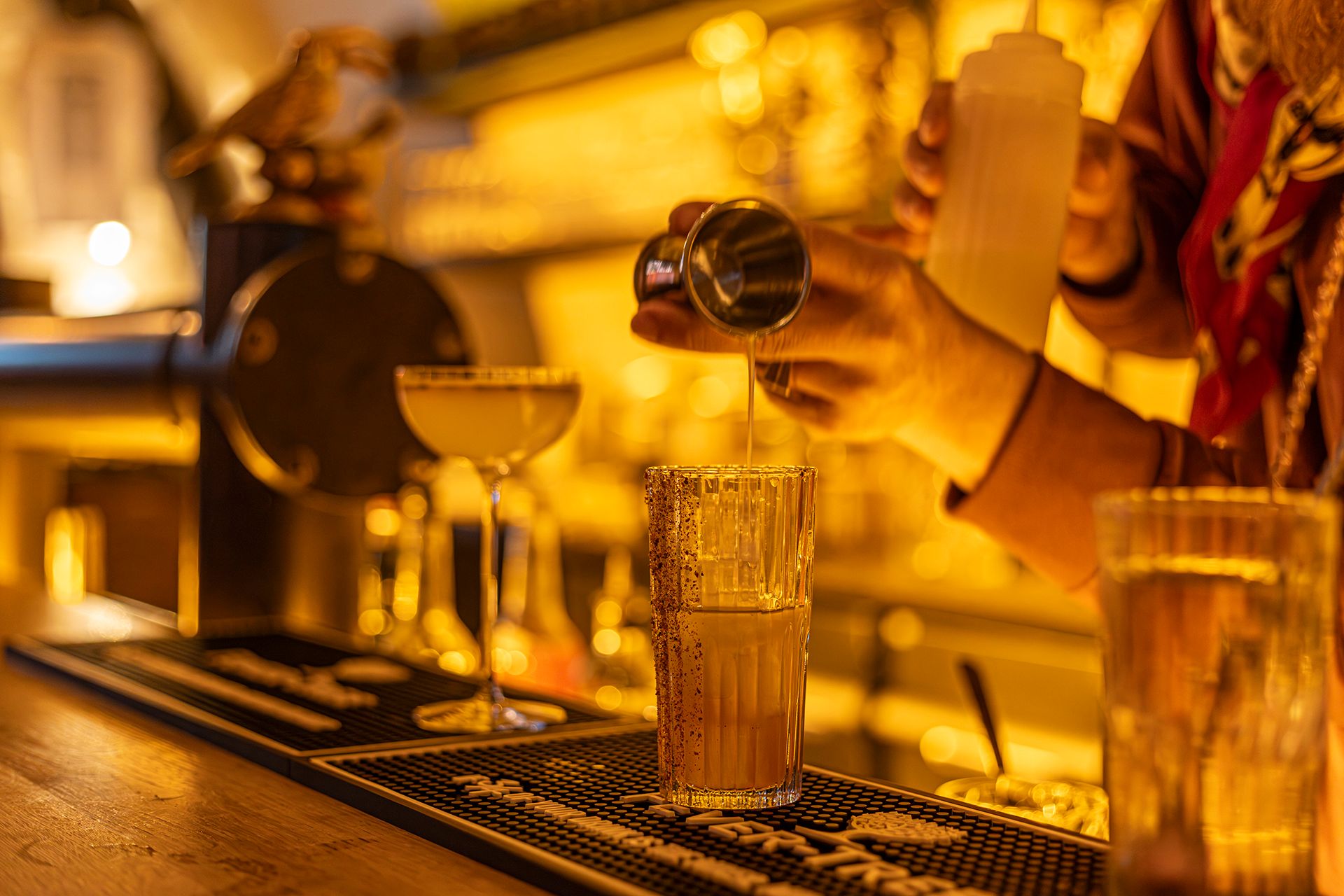 Ein Barkeeper mixt Cocktails in einer Bar mit warmem, gelbem Licht. Im Vordergrund stehen zwei Gläser, eines wird gerade befüllt.