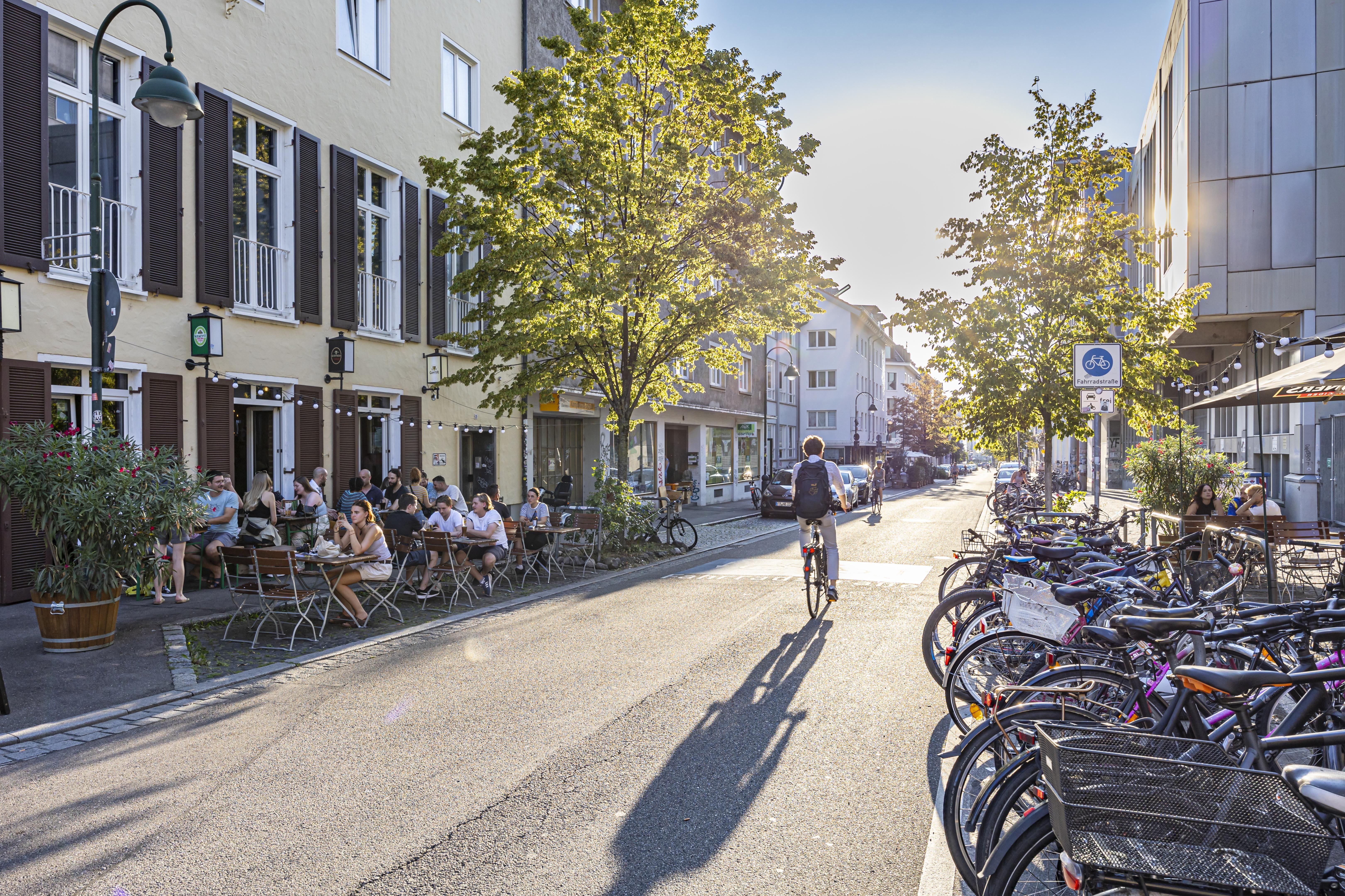 Straßenszene mit Radfahrern, Cafégästen und geparkten Fahrrädern bei Sonnenschein.